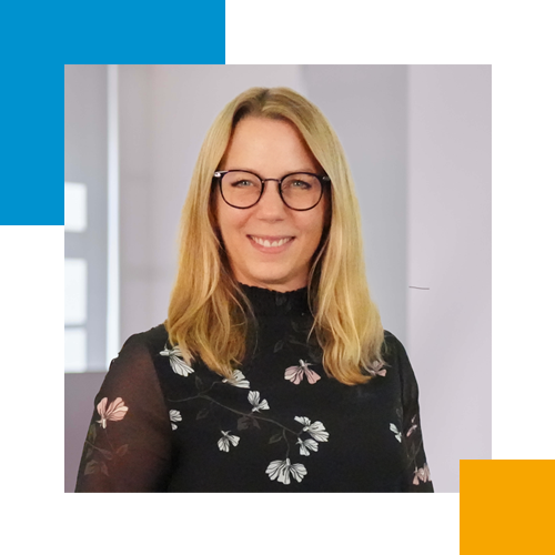 Eine junge Frau mit Brille und blonden Haaren, trägt ein schwarzes Kleid mit weißen und rosa Blumenmustern, lächelt und steht in einem hellen Raum.