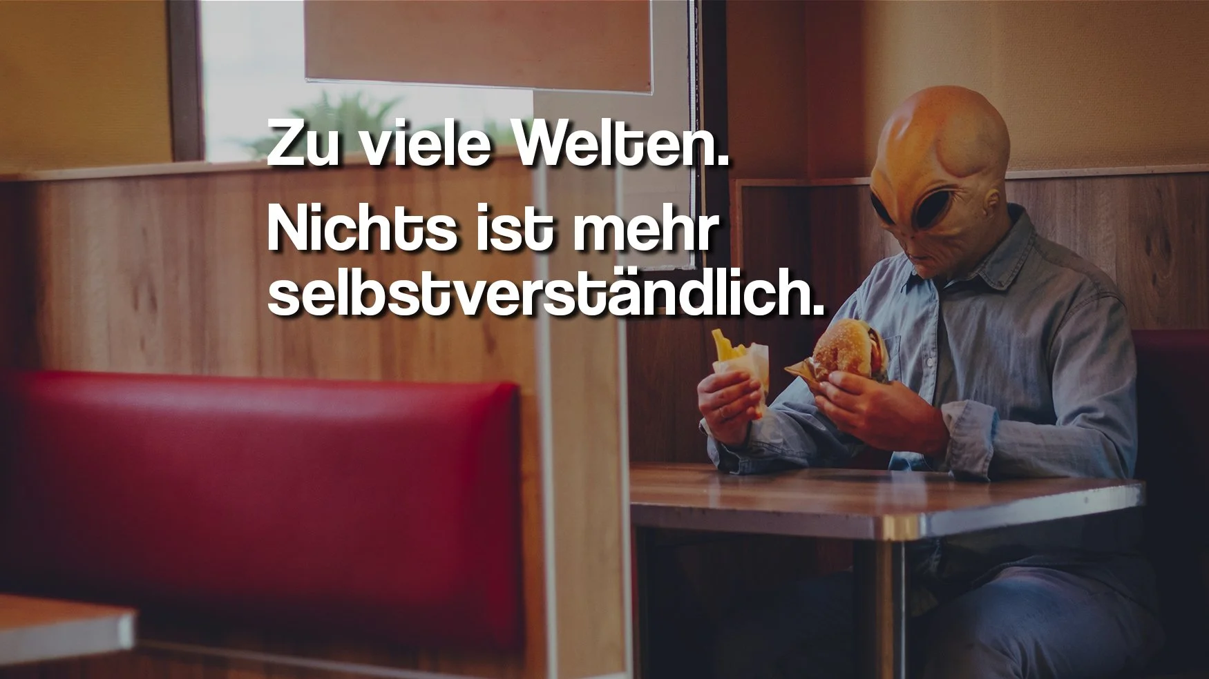 Eine Person mit einer Alienmaske sitzt in einem Fast-Food-Restaurant und isst einen Burger.