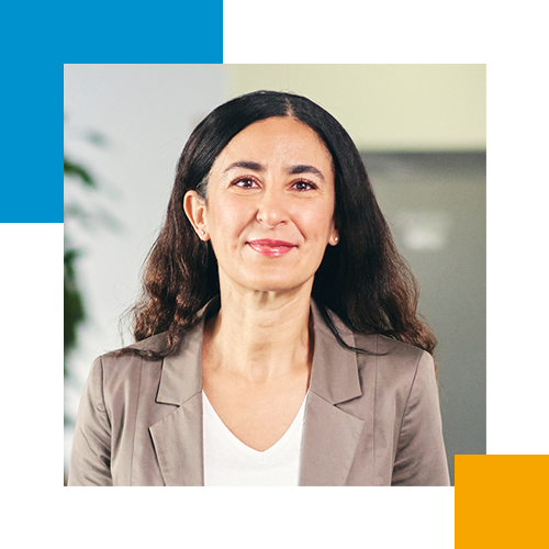 Eine Frau mit langem dunklem Haar, lächelnd, trägt einen grauen Blazer und ein weißes Top, im Büroumfeld.