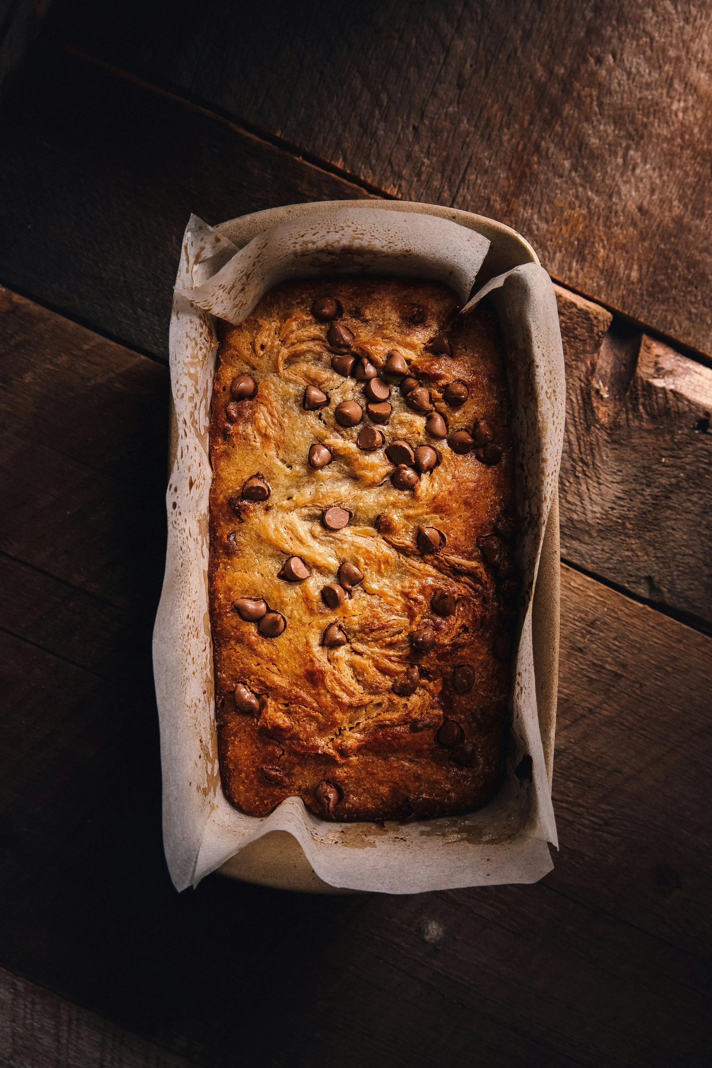 Banana bread with chocolate chips on top in a parchment-lined baking dish on a wooden surface.