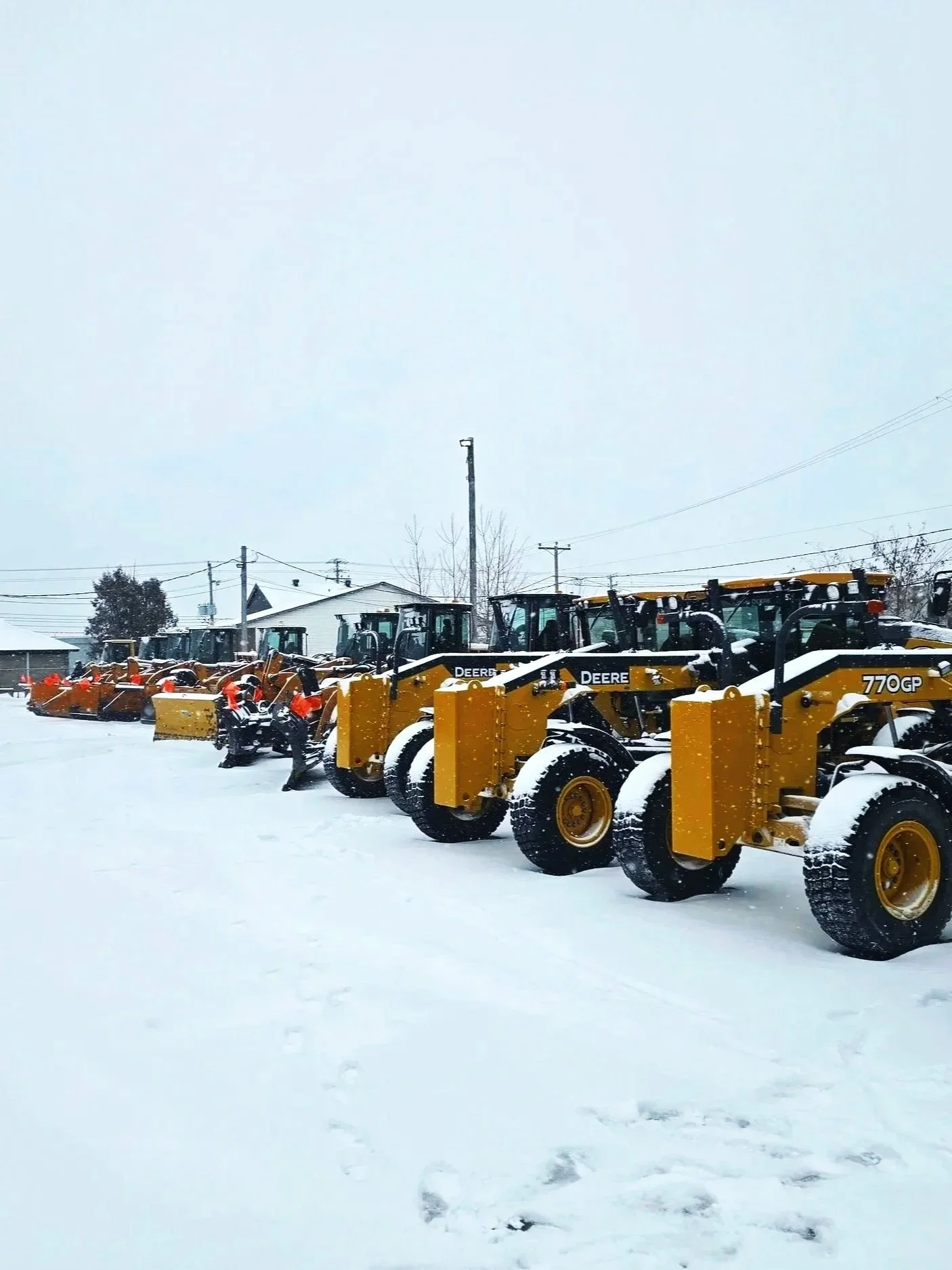 Une rangée de bulldozers jaunes stationnés dans une zone enneigée avec quelques personnes en vestes réfléchissantes en arrière-plan.