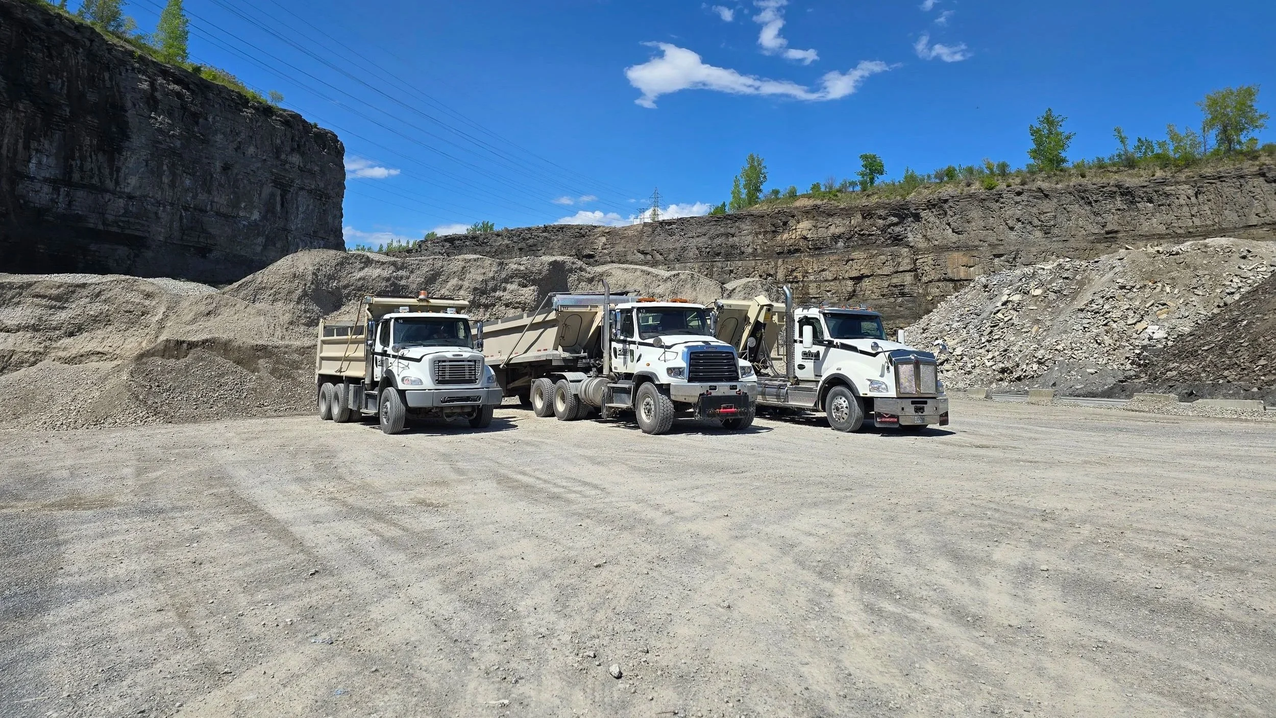 Trois camions de chantier stationnés dans un site de construction ou d'extraction de roche, avec un paysage rocheux en arrière-plan sous un ciel bleu avec quelques nuages.
