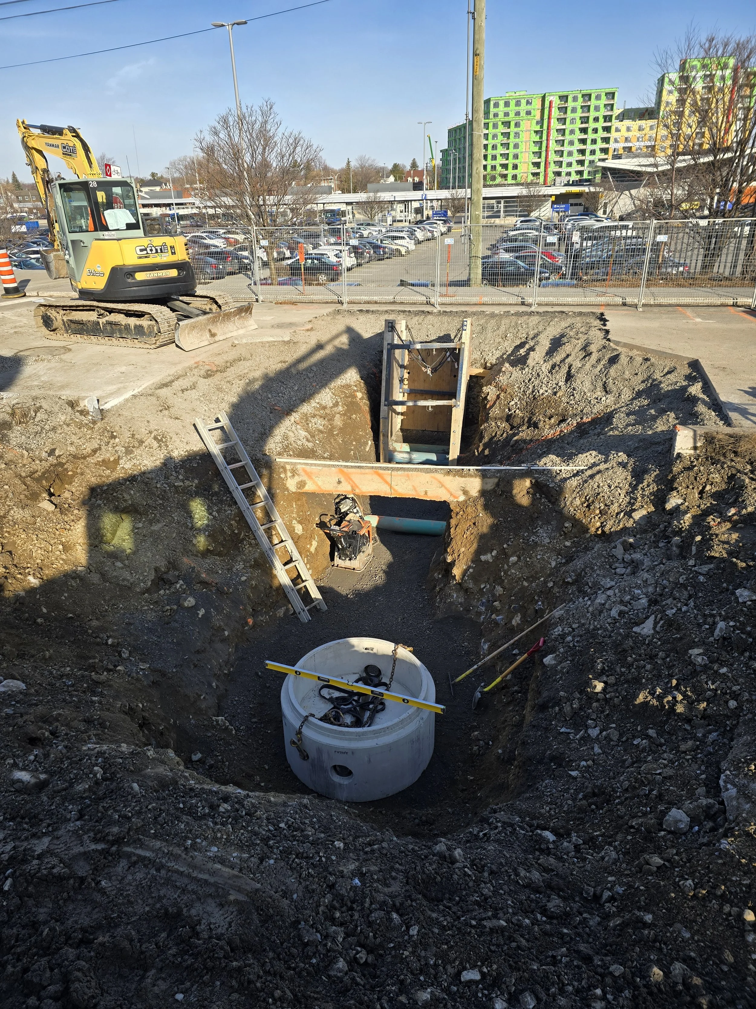 Travaux de construction avec une excavatrice et une fosse creusée dans le sol, contenant une grande cuve en béton et des tuyaux, dans un parking en milieu urbain.