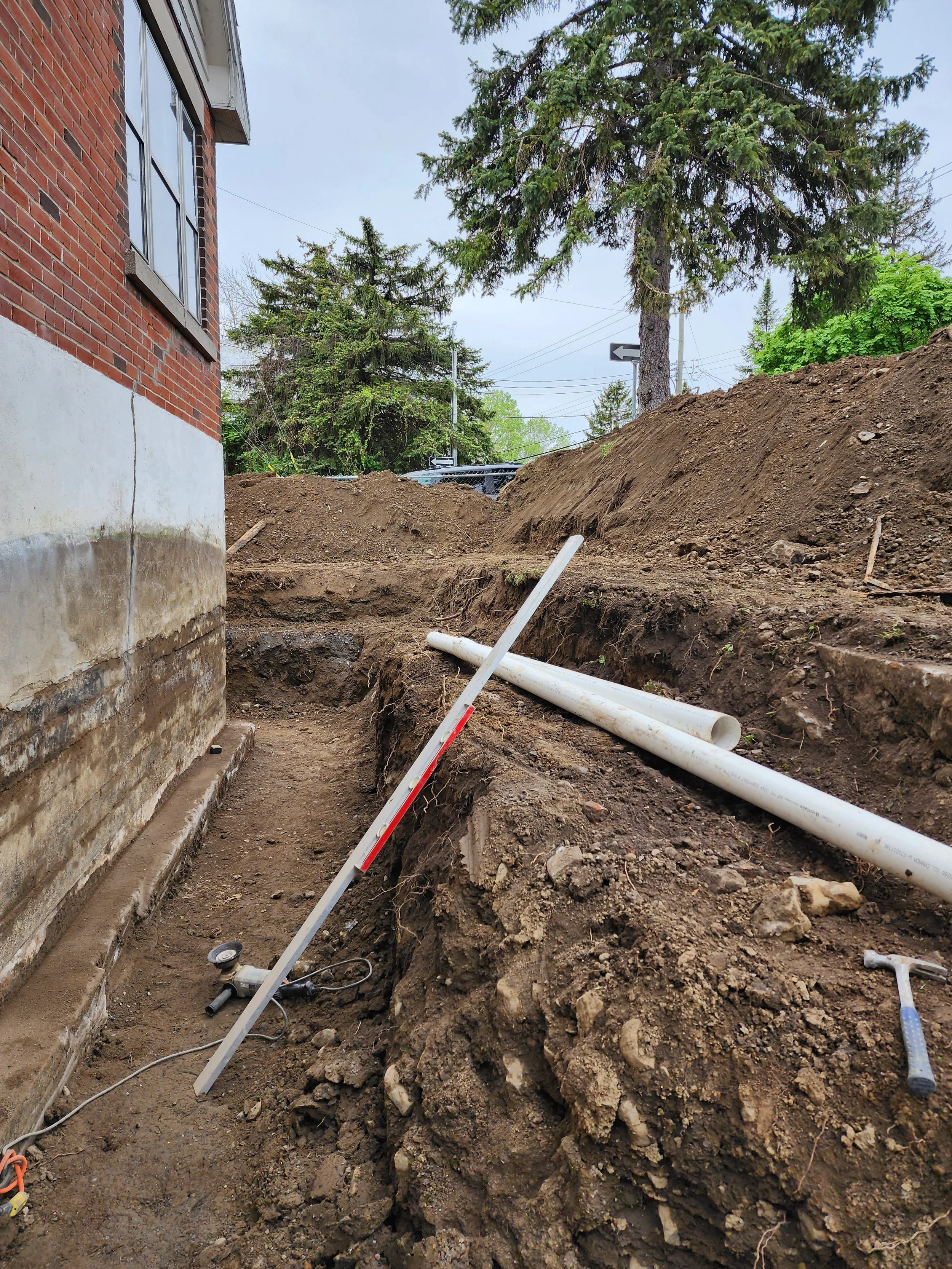 Travaux de plomberie en cours à côté d'une maison, avec des tuyaux en PVC noir et blanc, des outils et une excavation dans le sol.