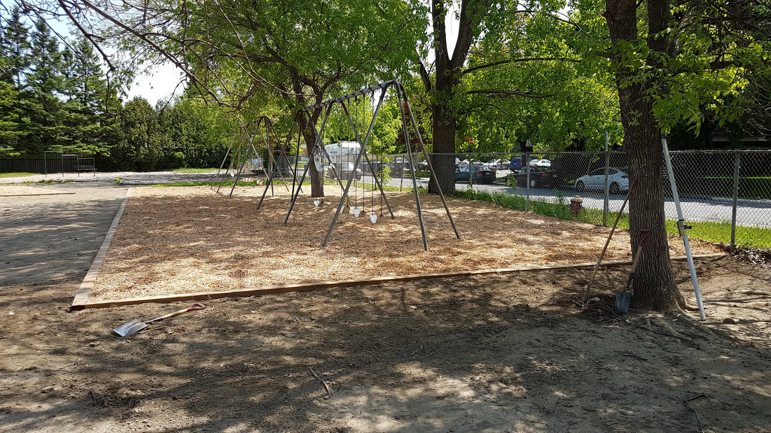 un terrain de jeu en train d'être aménagé avec des balançoires, installé sous un arbre, avec une clôture autour et une zone en gravier ou sciure ou copeaux de bois