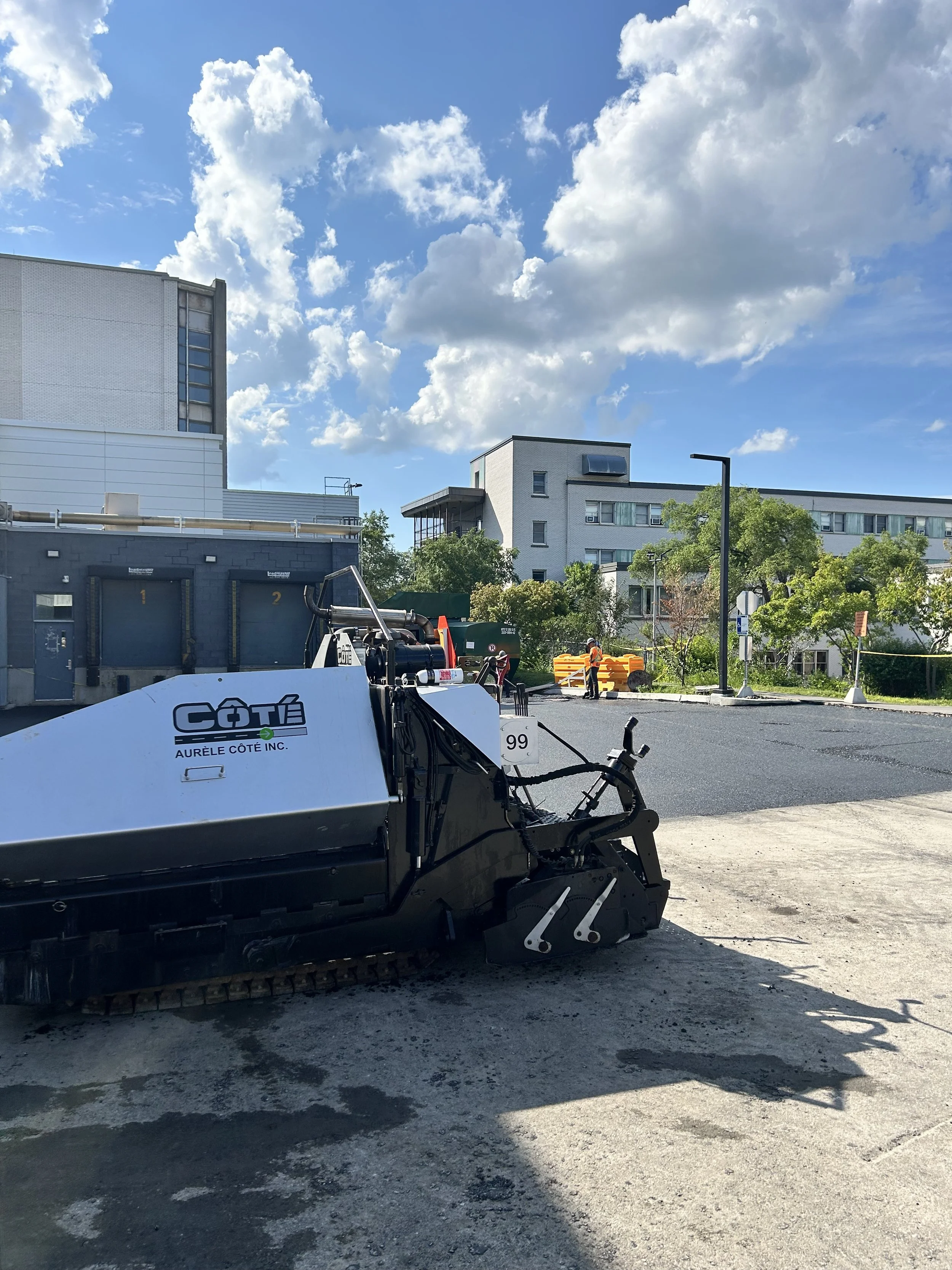 Une machine de construction de route qui compacte le bitume sur un parking en extérieur, avec des bâtiments modernes et des arbres en arrière-plan, sous un ciel bleu avec des nuages blancs.