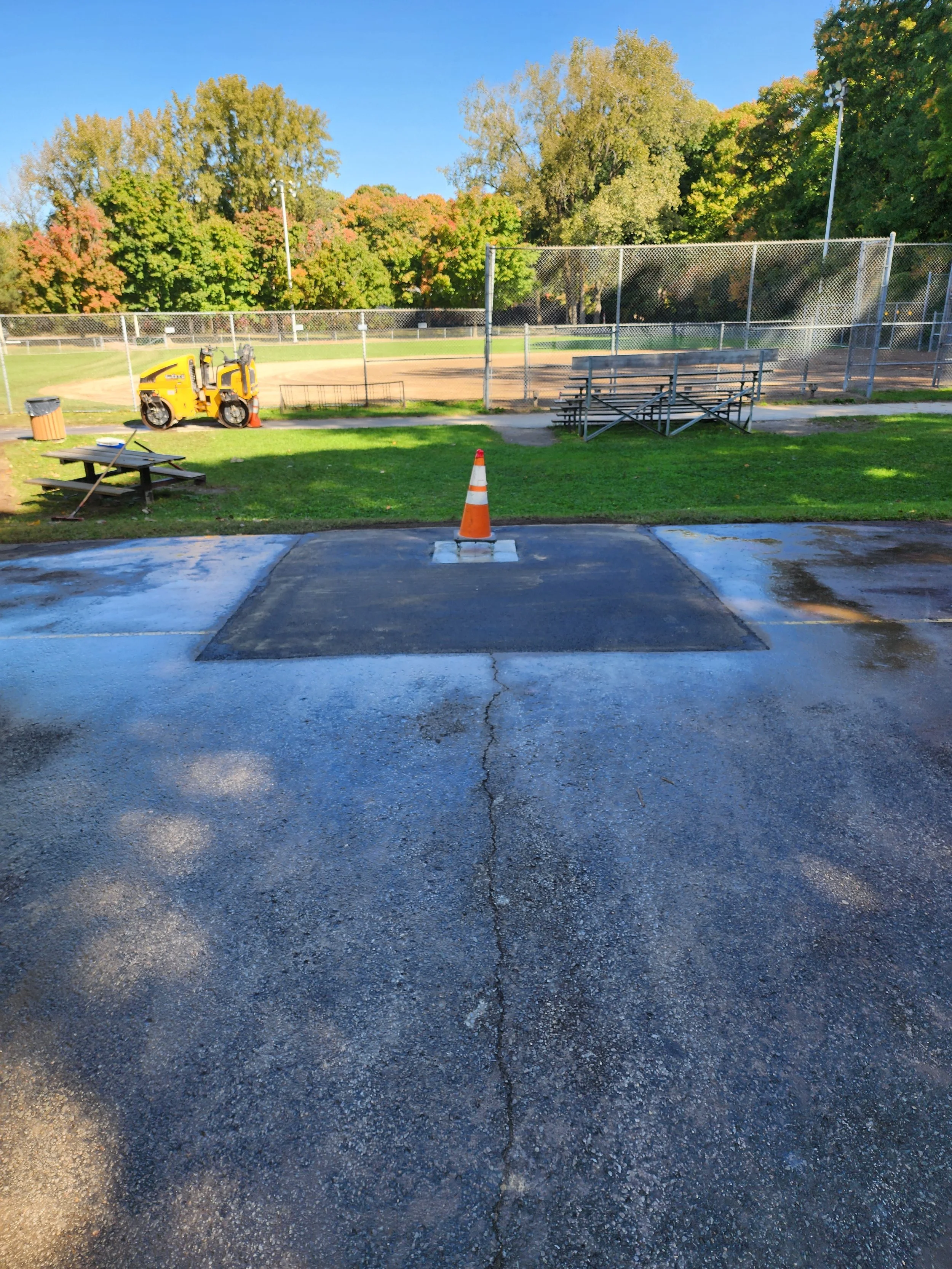 Zone de stationnement en cours de rénovation avec une barrière orange et un engin de nettoyage devant un terrain de baseball entouré d'une clôture, avec des arbres en arrière-plan sous un ciel bleu.