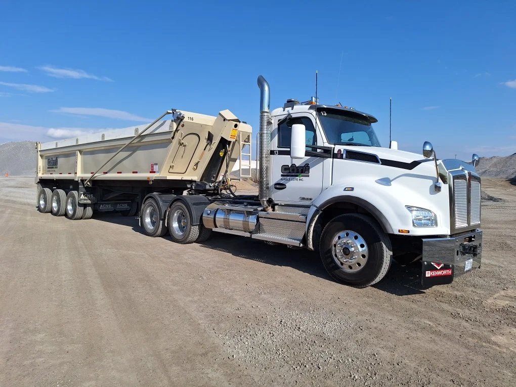 Un camion blanc avec une remorque beige dans un site de construction ou de mine, sur un sol en terre, avec un ciel bleu clair avec quelques nuages.