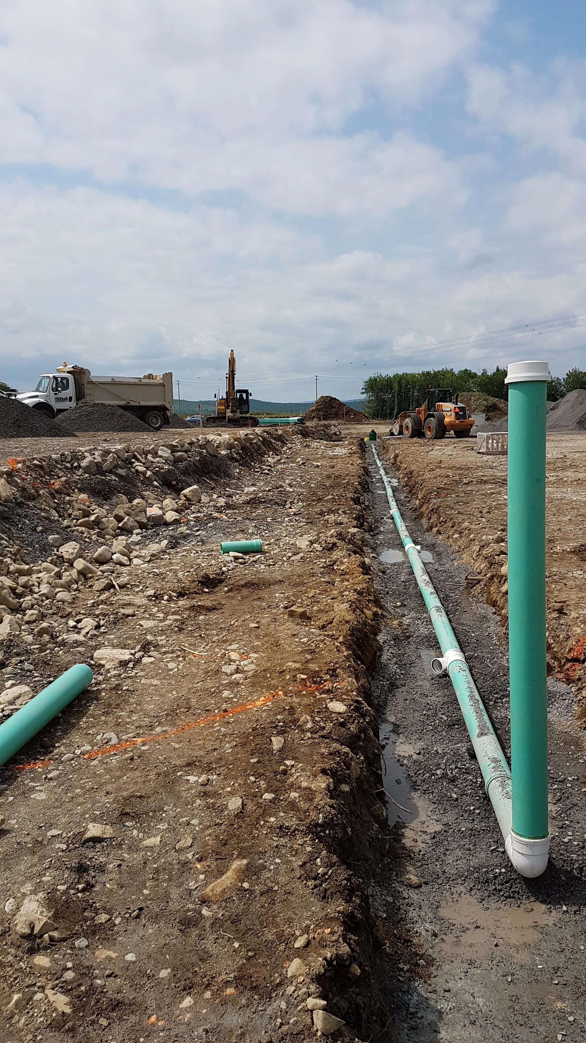 Travaux de construction avec des tuyaux en PVC turquoise installés dans une tranchée, engins de chantier en arrière-plan, ciel nuageux.