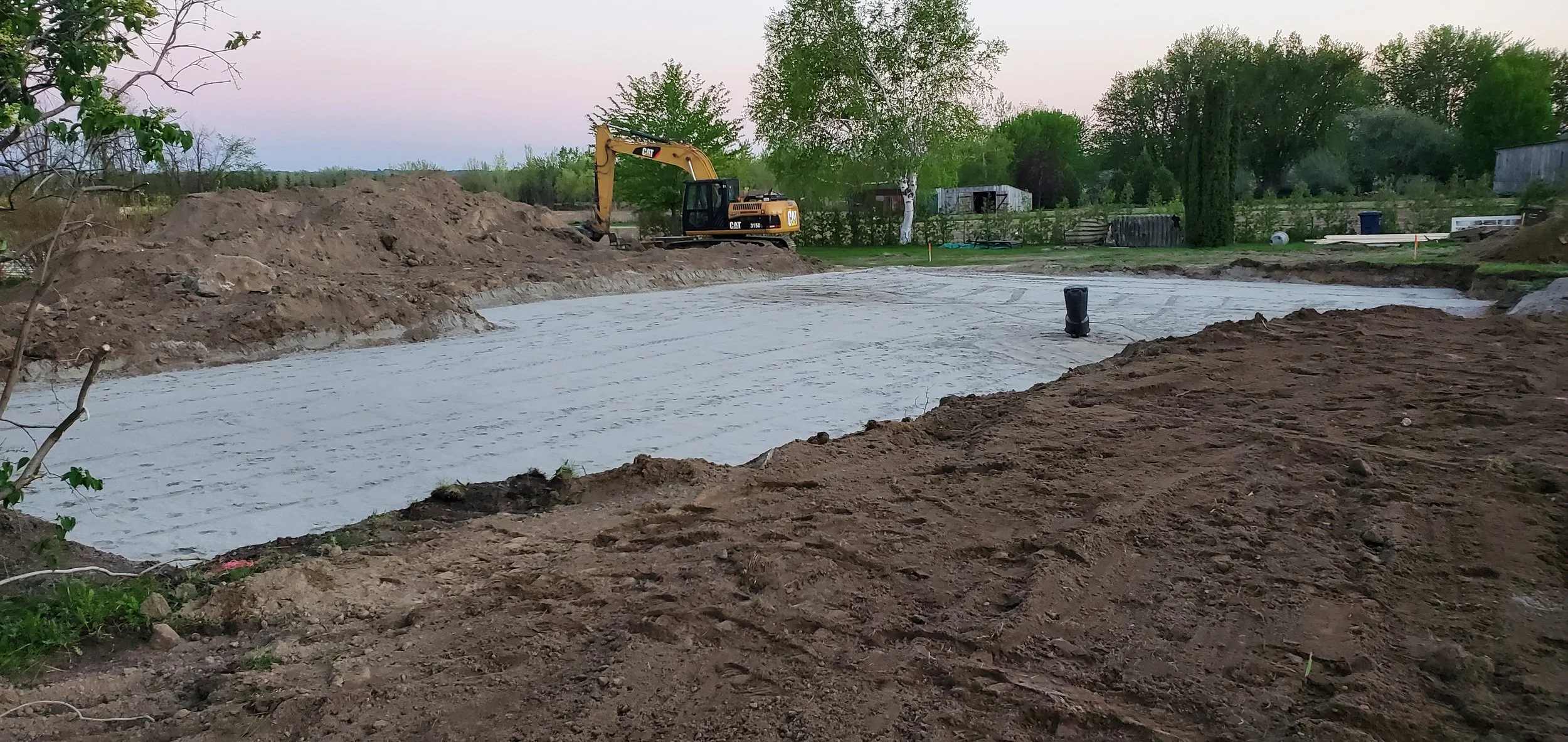 Chantier avec une excavatrice jaune en arrière-plan, sol en cours de préparation, un revêtement blanc de béton ou de plastique recouvre un terrain, entouré d'arbres et de végétation.