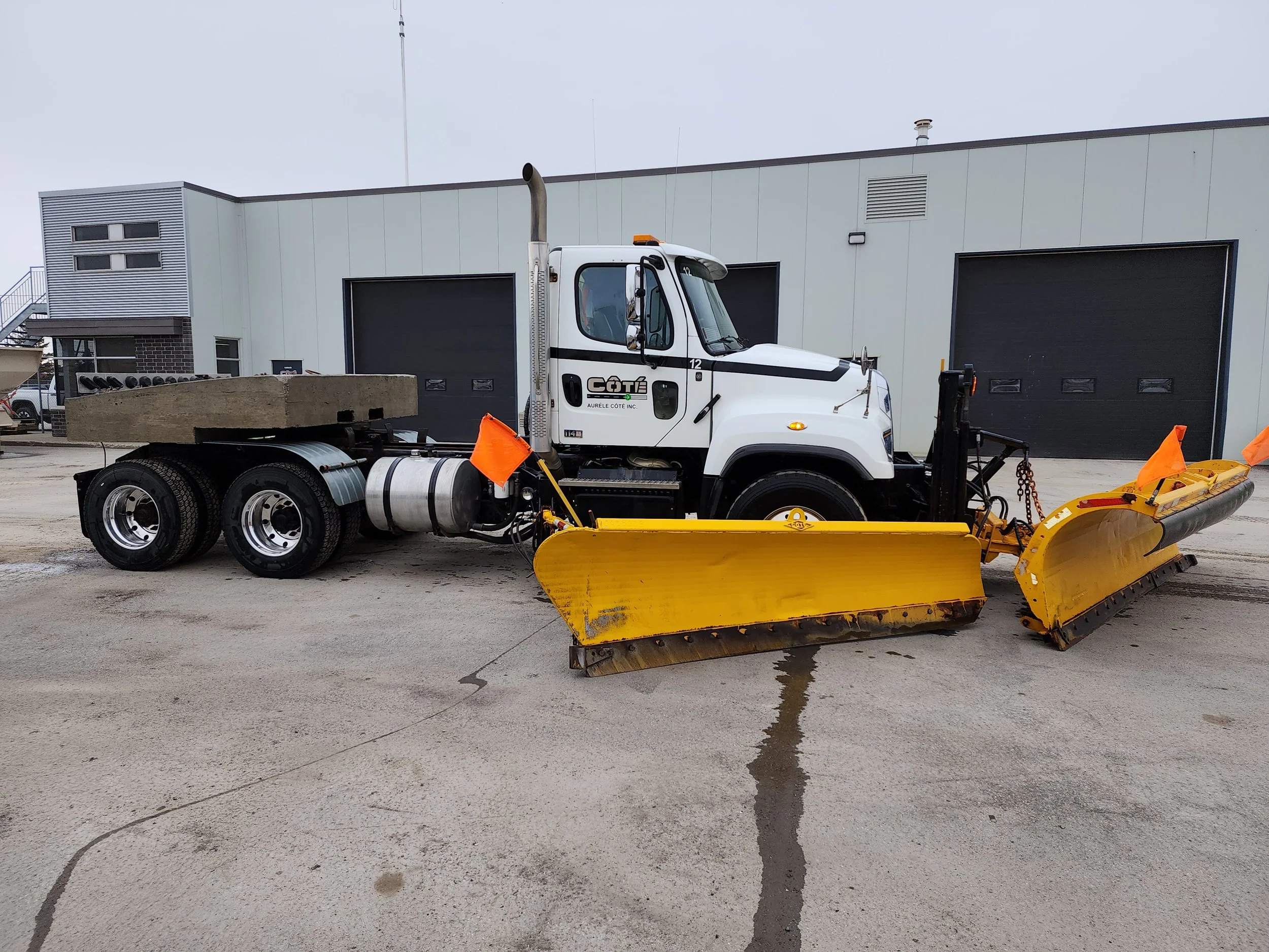 Camion avec une lame de déneigement jaune attachée à l'avant, stationné sur une surface en béton devant un bâtiment industriel.