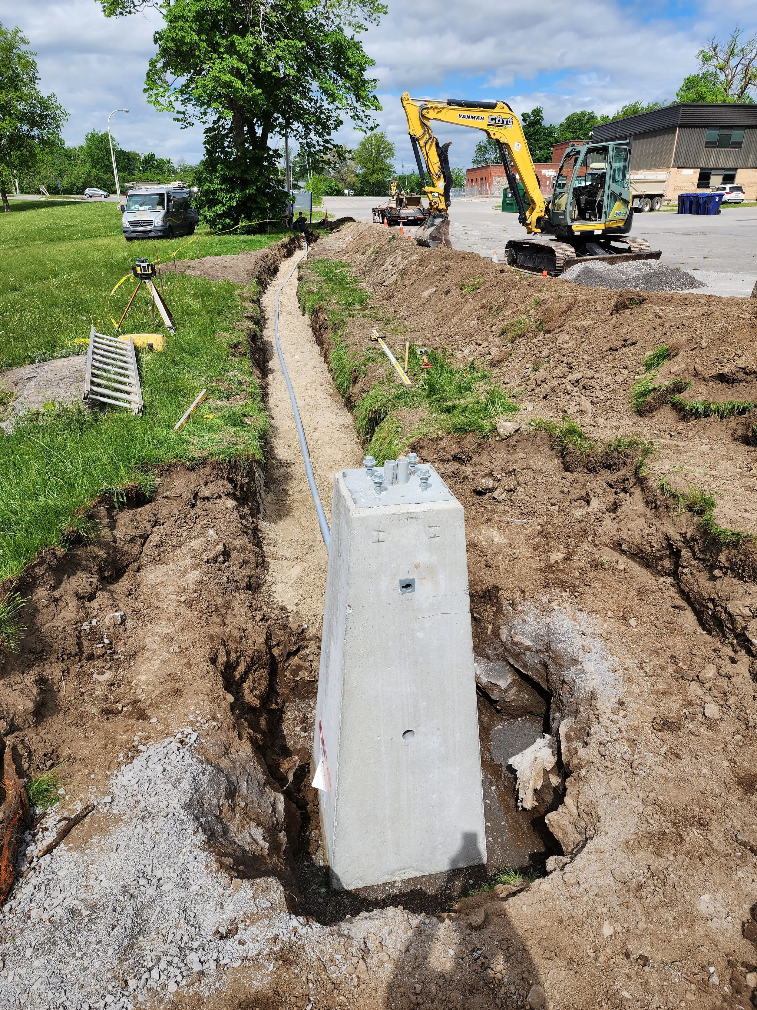 Travail de construction avec une tranchée pour des câbles ou des tuyaux, une pelle mécanique, et des repères de chantier dans un espace extérieur.