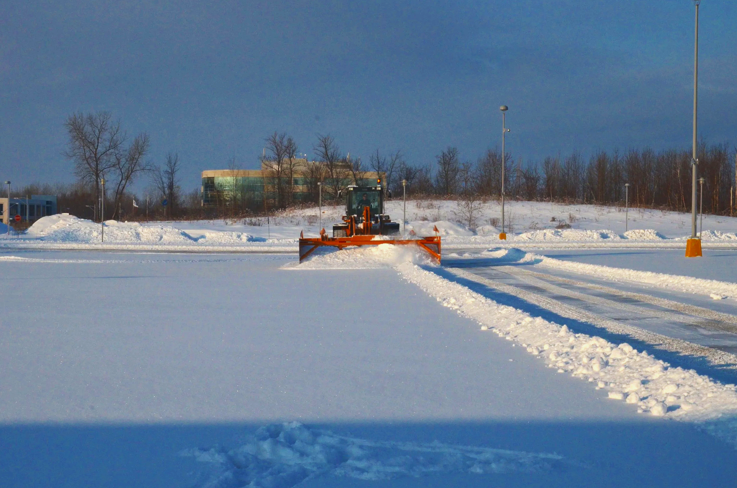 Un engin de déneigement orange en train de pousser la neige sur une surface enneigée, avec des lampadaires et des arbres en arrière-plan, par temps ensoleillé.