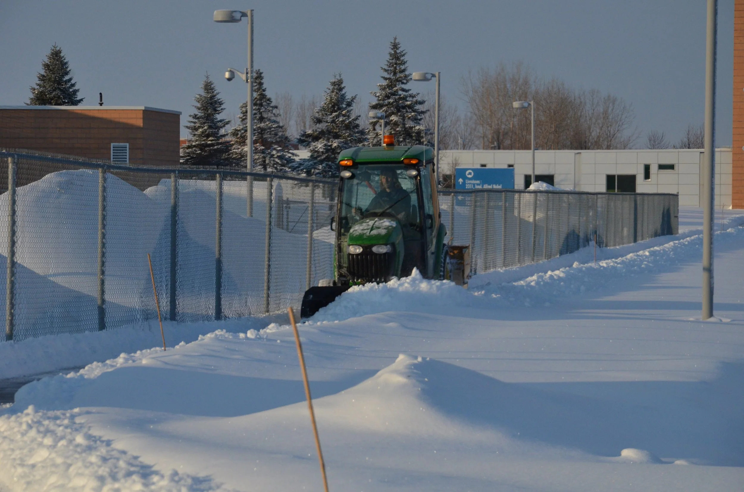 Un tracteur vert qui déblaye la neige sur un chemin pavé entouré de clôtures en métal, sous un ciel clair d'hiver.