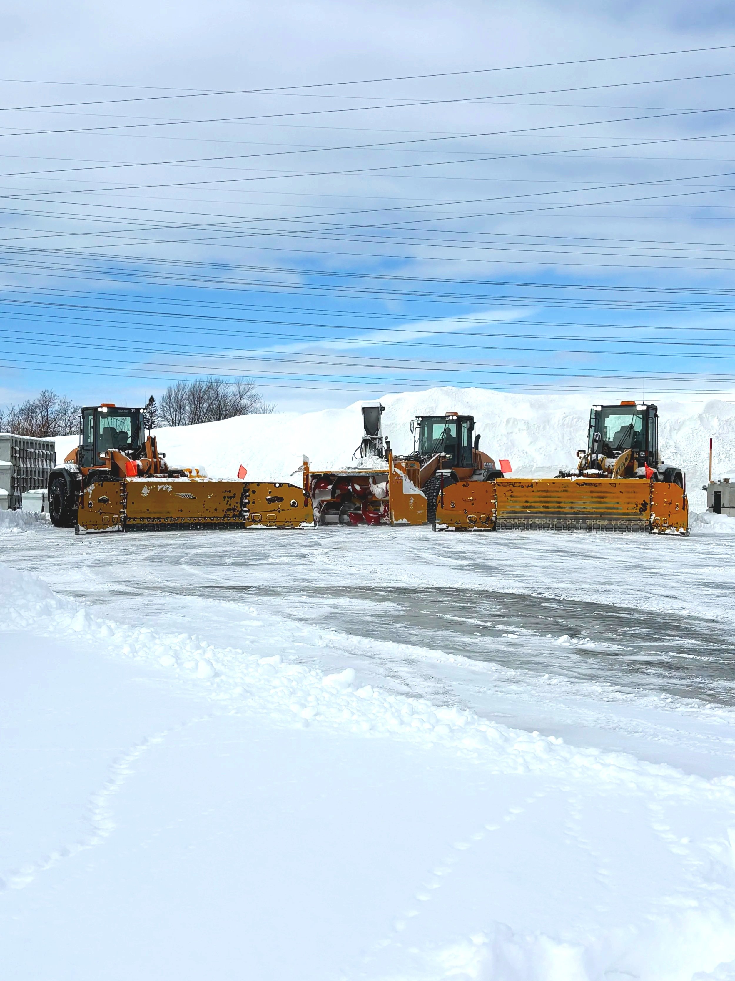 Trois véhicules de déneigement orange déblayent la neige sur une surface givrée dans un environnement enneigé, avec des câbles électriques au-dessus et un ciel partiellement nuageux.