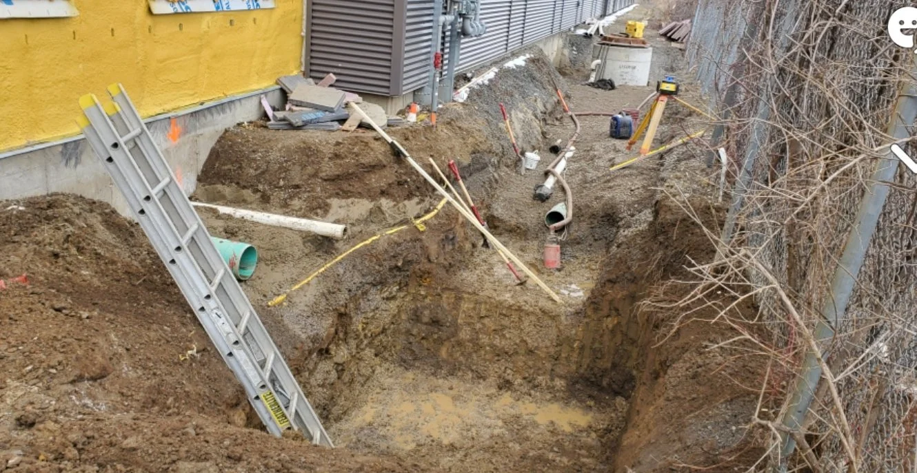 Travaux de excavation avec outils et équipements de chantier, tuyaux apparents, et terrain en travaux, à proximité d'une clôture métallique et d'un bâtiment.