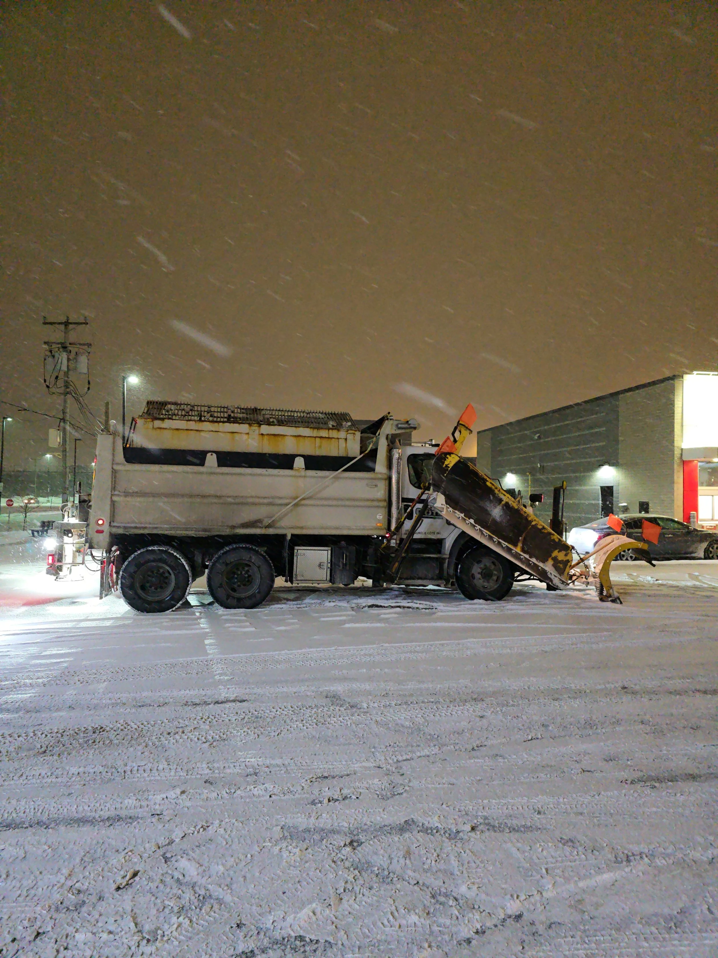 Un camion de déneigement stationné dans une zone urbaine enneigée avec un homme en train de nettoyer la neige. Il y a plusieurs voitures en arrière-plan, un bâtiment moderniste et des lampadaires illuminant la scène nocturne.