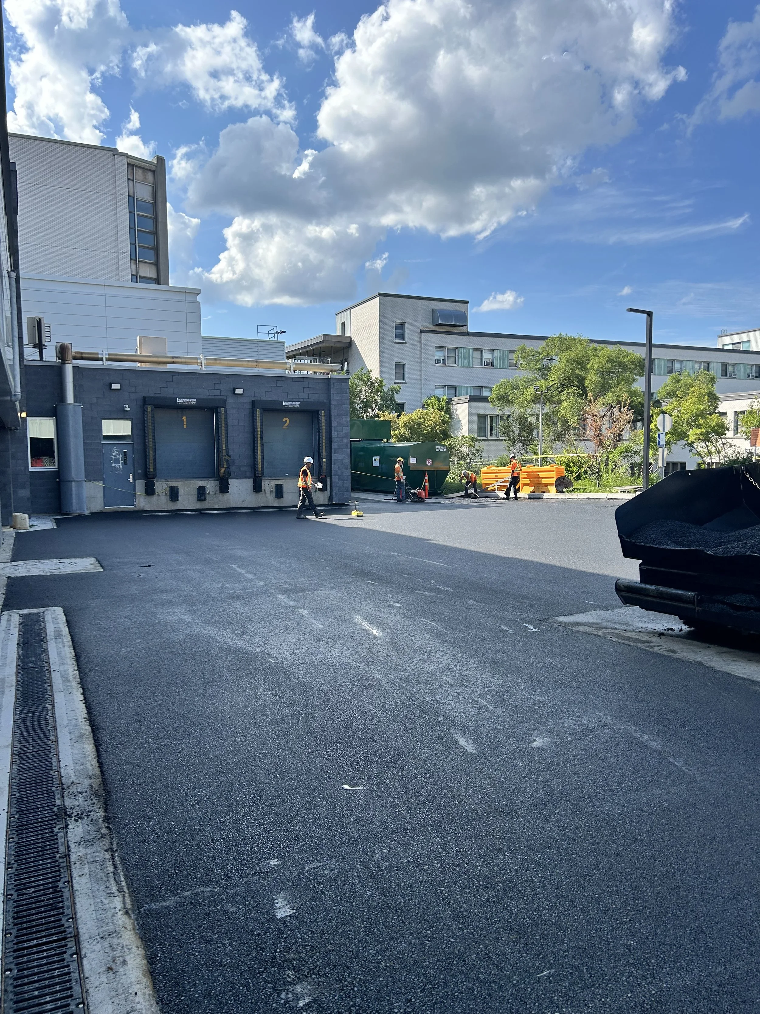 Travaux routiers avec ouvriers en sécurité, route fraîchement asphaltée, bâtiments modernes et ciel nuageux en arrière-plan.