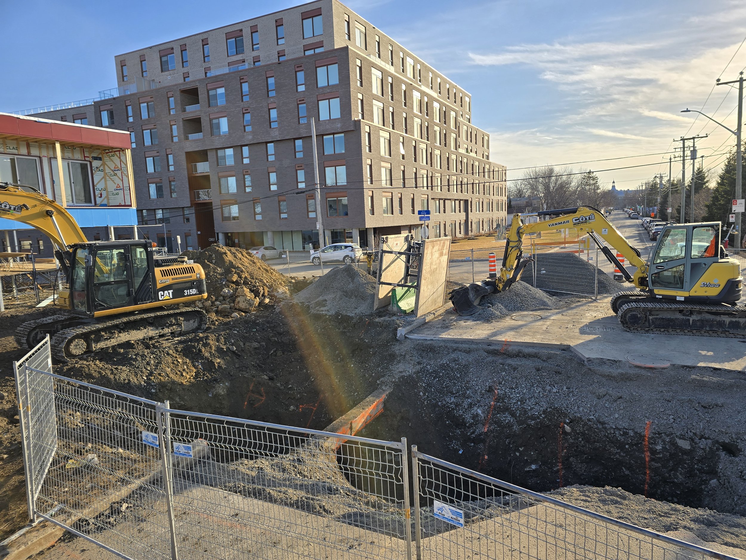 Chantier de construction avec deux excavatrices jaunes, une grande excavation, et un bâtiment moderne en arrière-plan, sous un ciel ensoleillé.