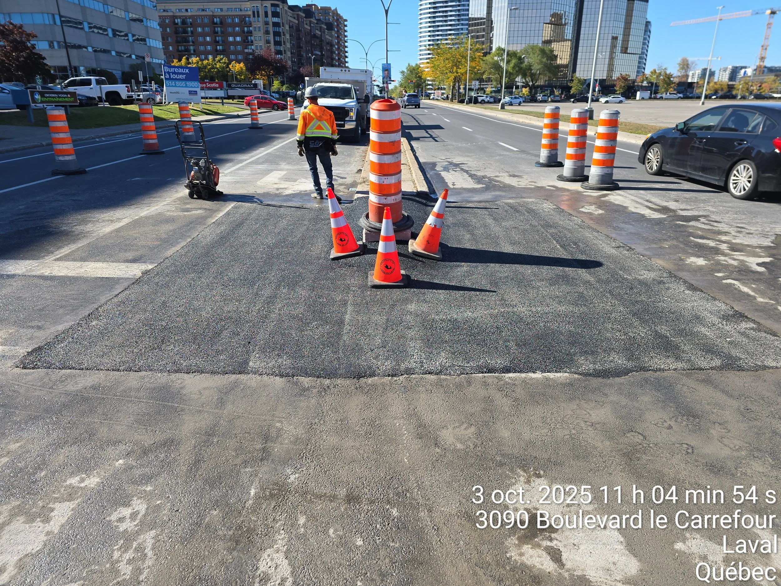 Travaux de voirie avec des cônes orange et un homme en veste de sécurité travaillant sur une route en milieu urbain à Laval, Québec.