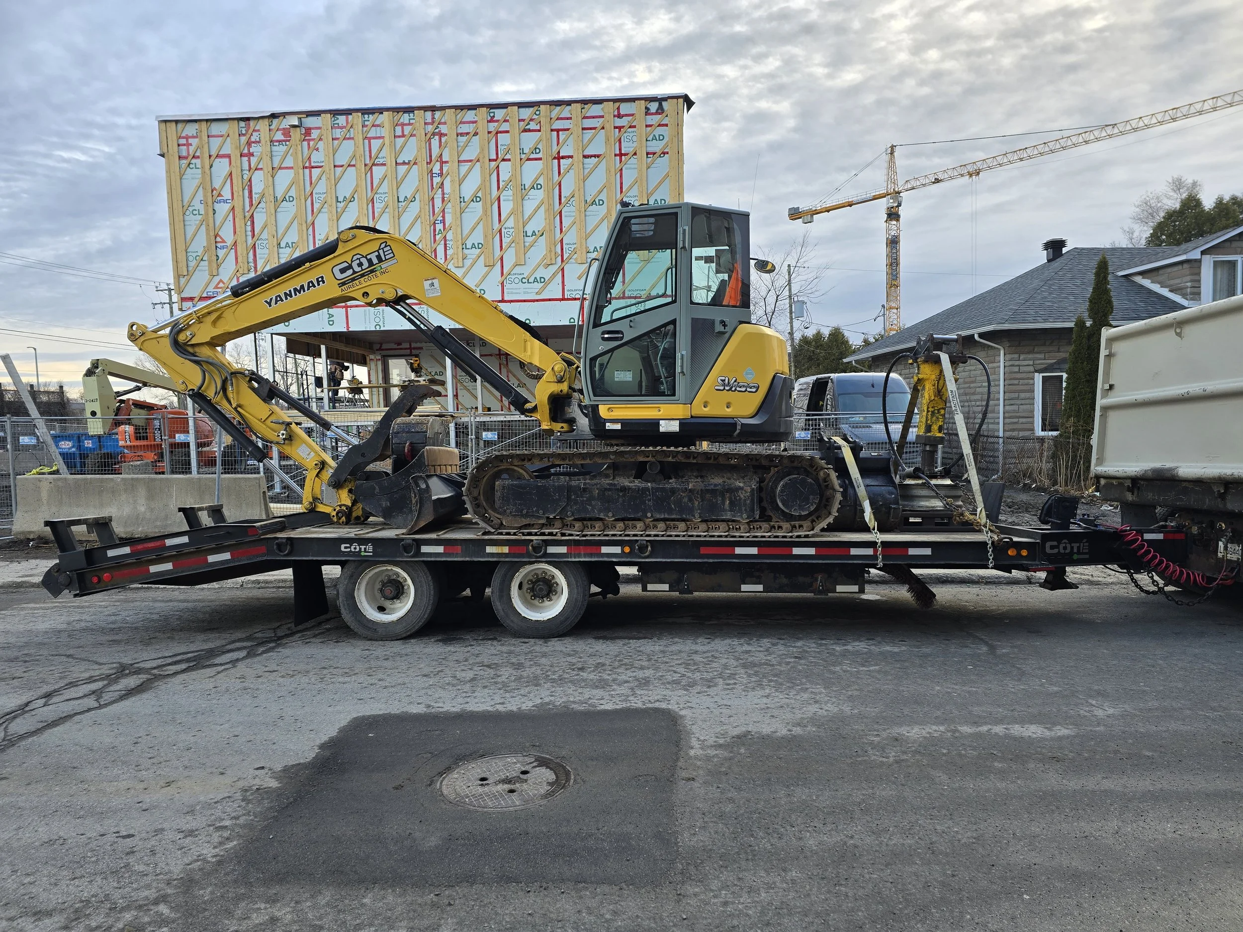 Mini excavatrice jaune sur une remorque au chantier avec une maison en construction en arrière-plan.