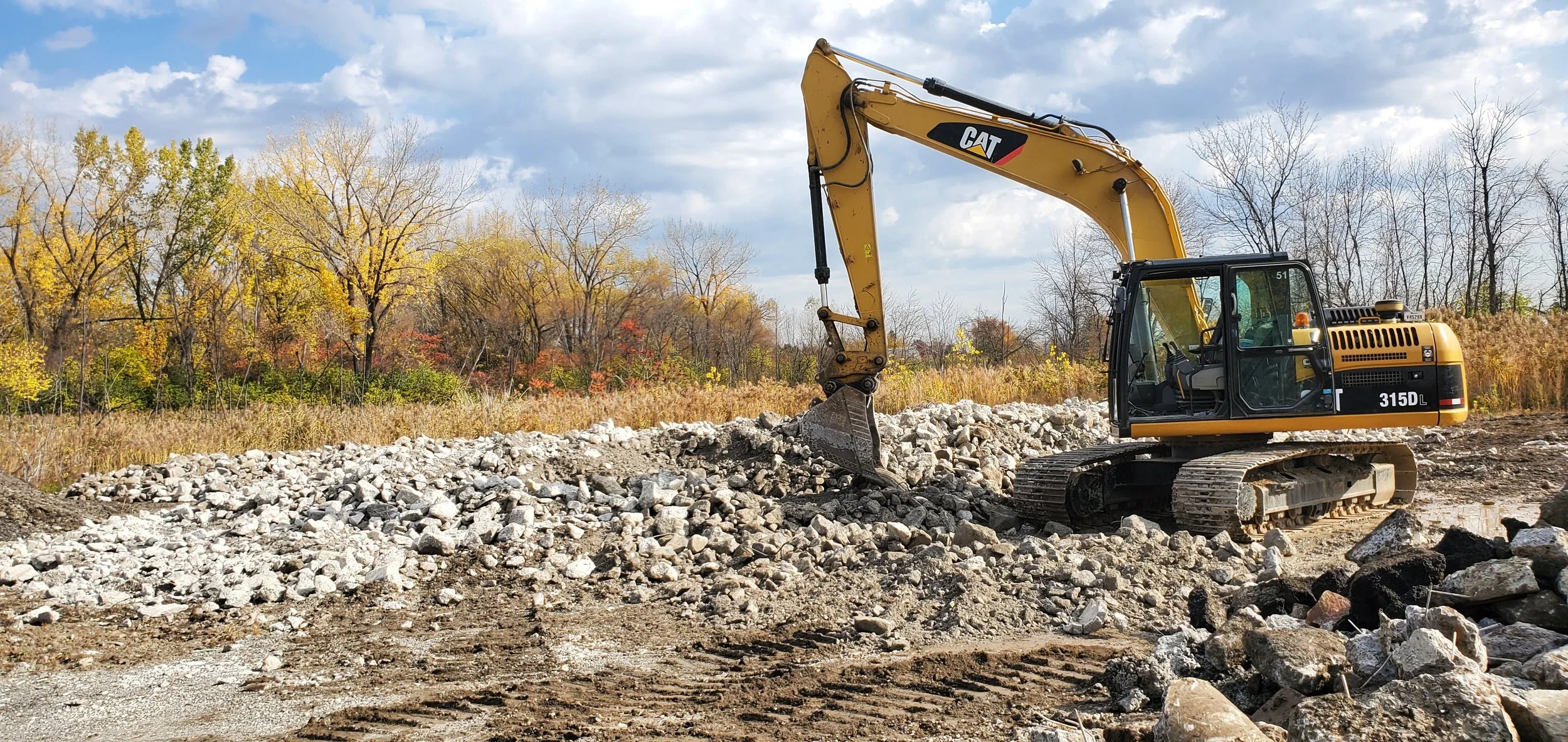 Mini excavatrice jaune de marque CAT en action sur un chantier de construction avec une couche de roches et de terre, arrière-plan de arbres aux couleurs automnales et ciel partiellement nuageux.