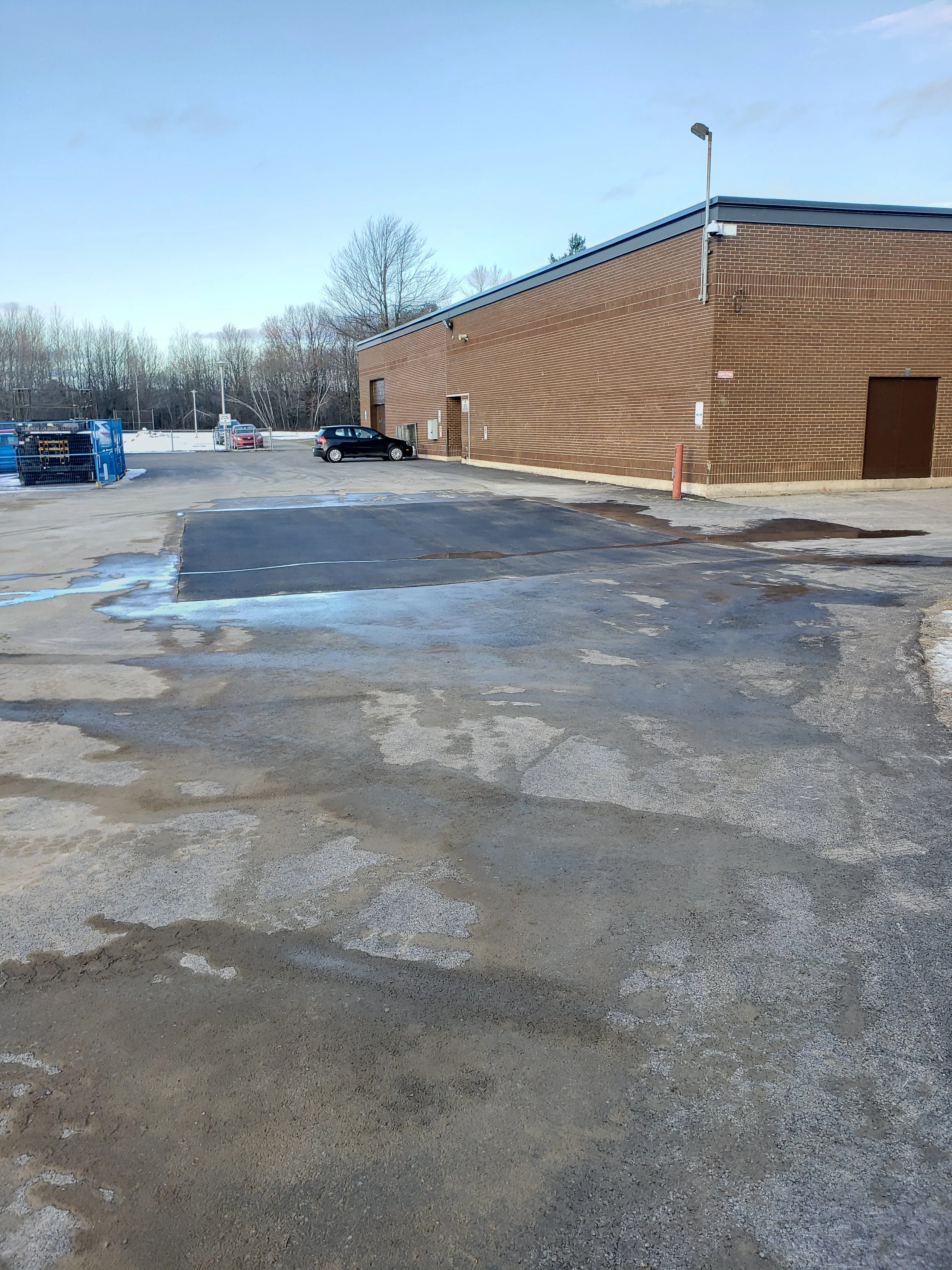 Un parking vide avec quelques voitures à l'arrière-plan, une grande bâtisse en briques, des flaques d'eau sur le sol, et un ciel partiellement nuageux.