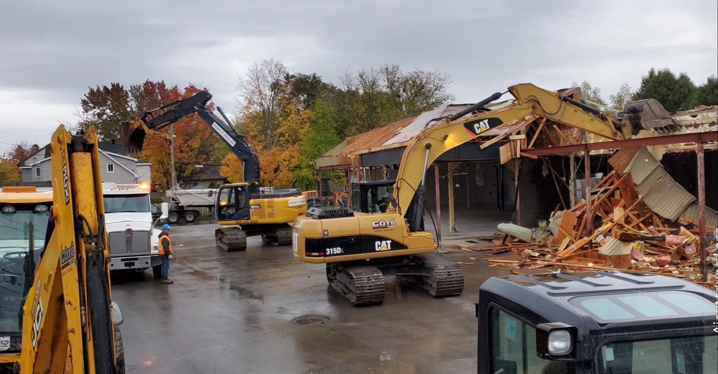 Des engins de construction détruissent un bâtiment en structure en bois avec des débris éparpillés, sous un ciel nuageux, dans un environnement résidentiel avec des maisons en arrière-plan.