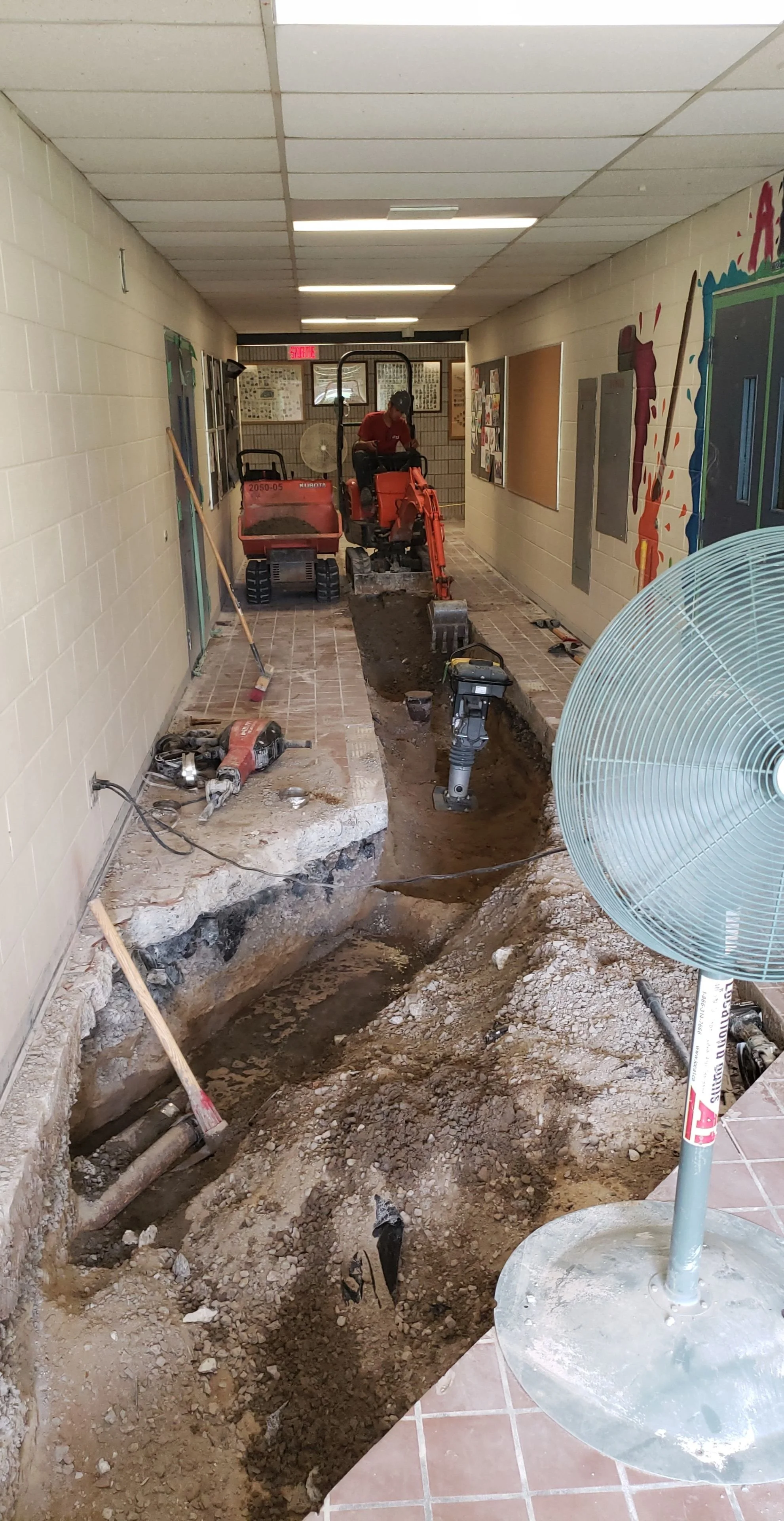 Travaux de rénovation dans un couloir scolaire, avec une tranchée creusée, des outils et une mini-pelle.