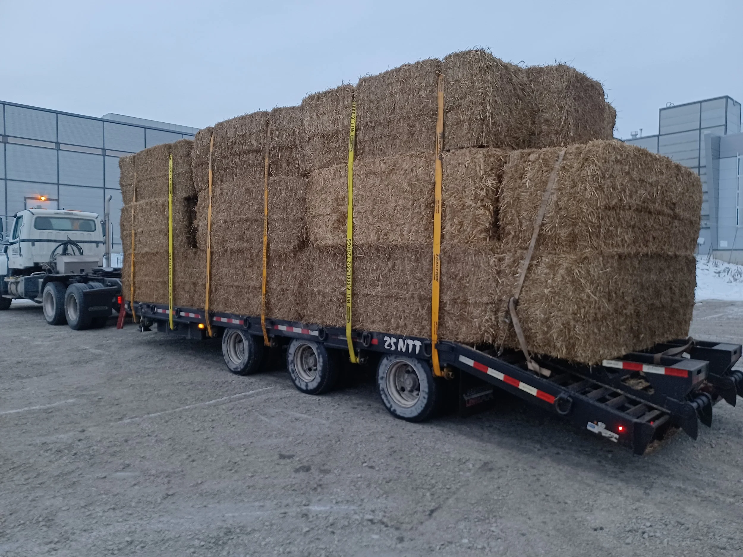 Un camion transportant de la paille empilée dans des balles rectangulaires, garé dans un parking en extérieur avec un bâtiment industriel en arrière-plan.