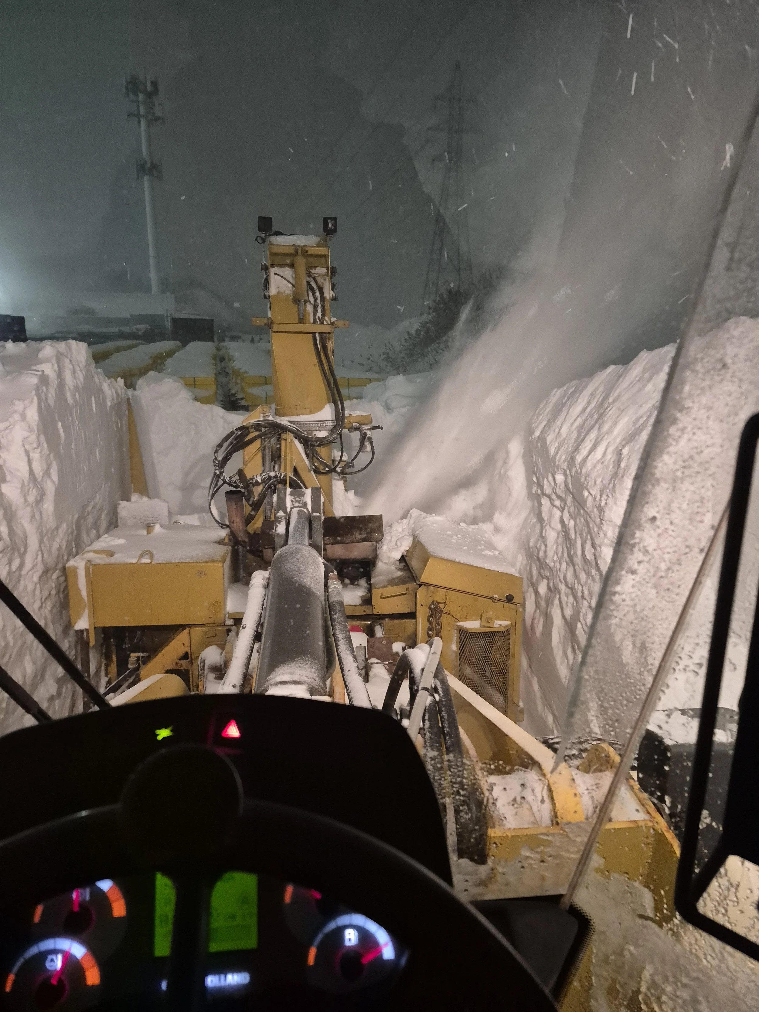Vue depuis l'intérieur d'un véhicule en marche, montrant une machine de déneigement en action en pleine tempête de neige la nuit.