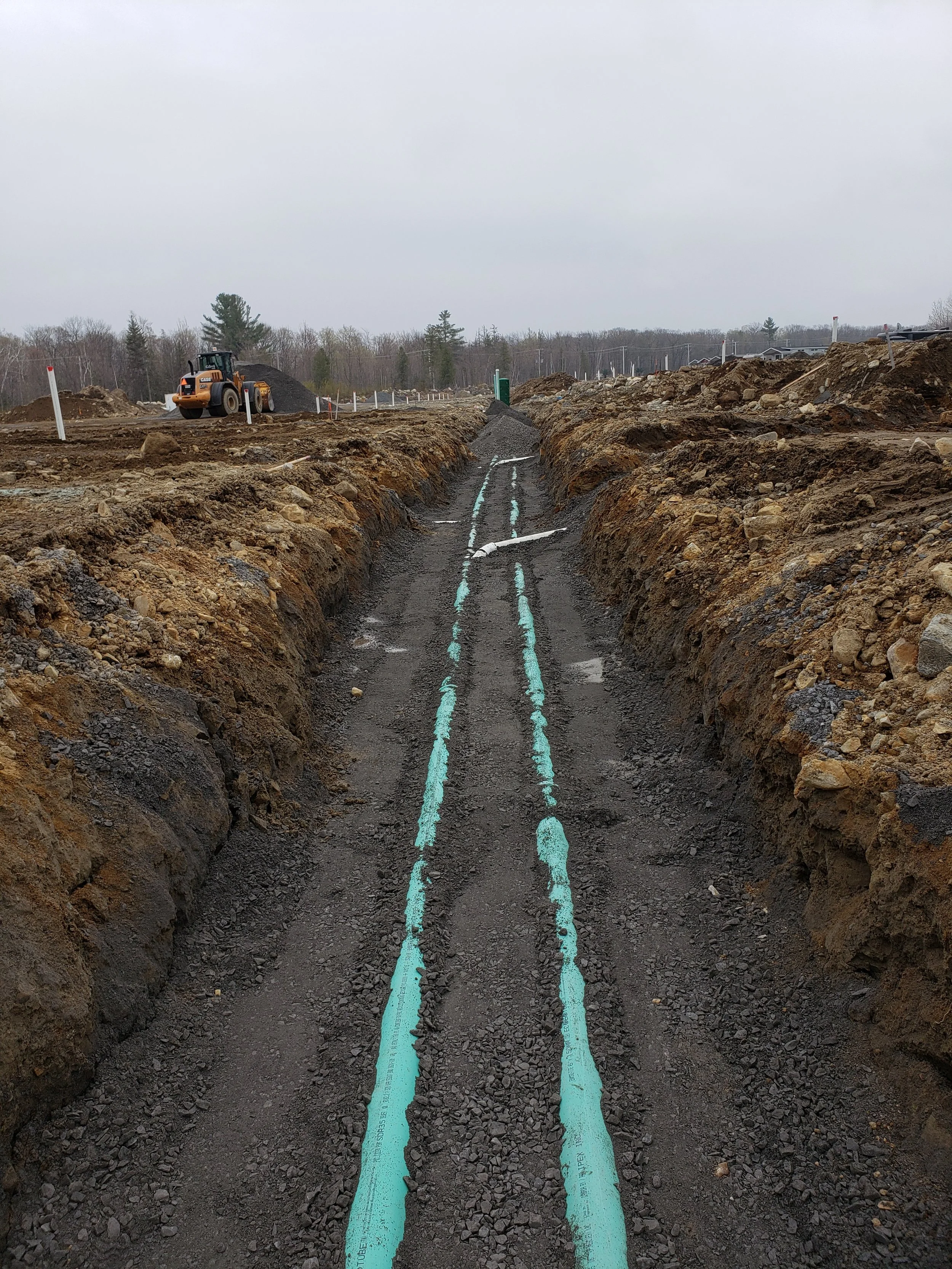 Une tranchée en construction avec des lignes de canalisation peintes en bleu, un engin de chantier à gauche, et un paysage de terrain accidenté avec des arbres en arrière-plan.