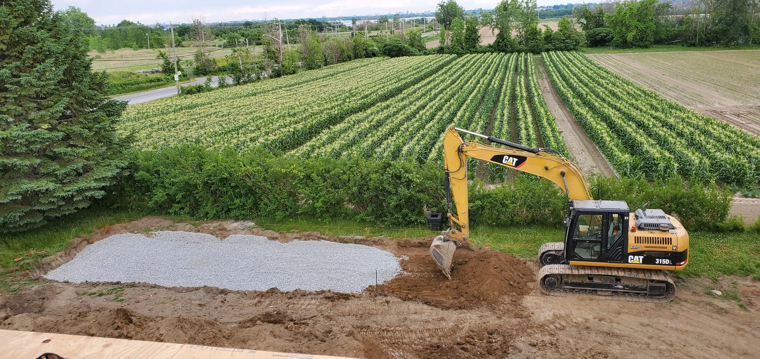 Mini excavatrice jaune en plein travail dans un jardin potager, préparant un espace avec du gravier, avec des champs cultivés en arrière-plan.