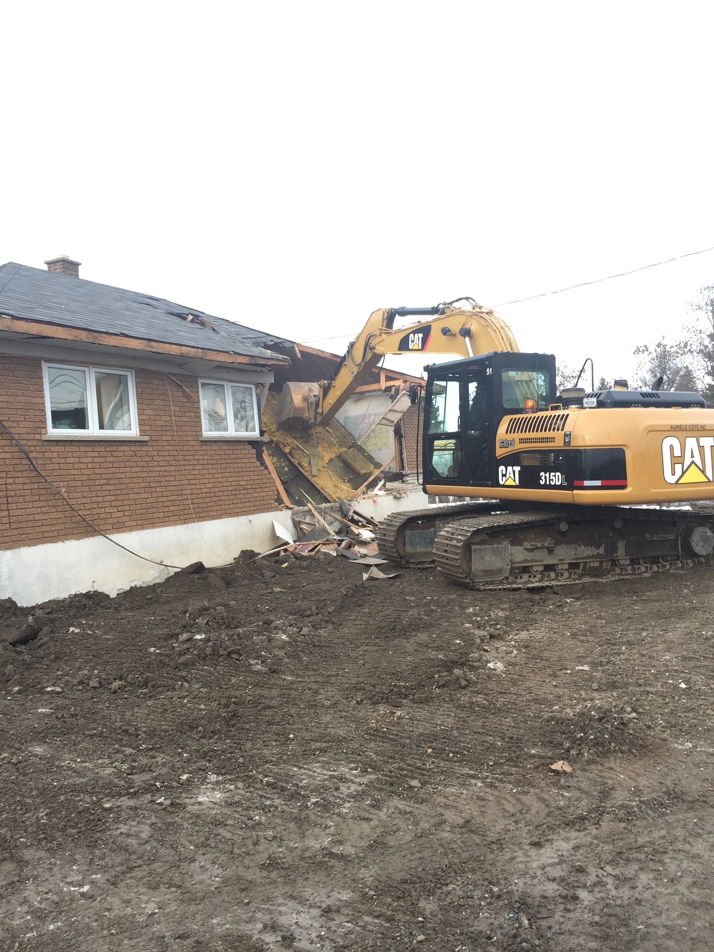 Une pelleteuse Caterpillar en train de détruire une partie d'une maison en briques, avec du terrain excavé en premier plan.