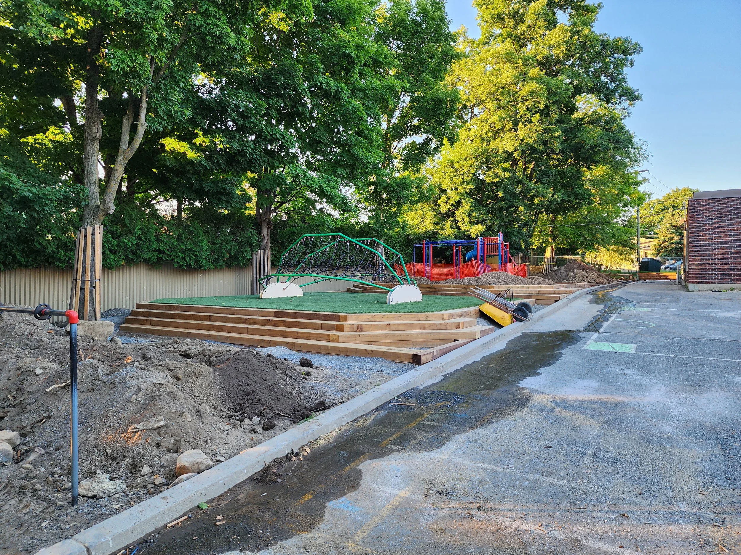 Terrain de jeu en construction avec une plateforme en bois, une arche de piratage, un terrain multisport avec un sol enrobé, et des arbres en arrière-plan.