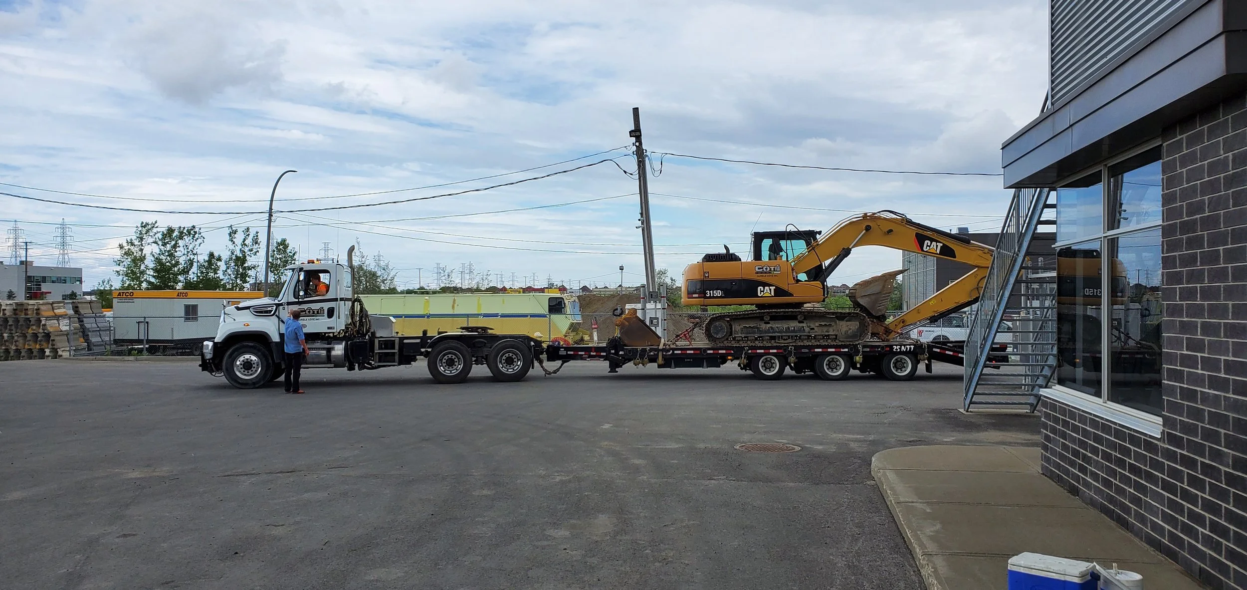 Un camion de transport transporte une petite excavatrice jaune sur son plateau dans un parking extérieur, près d'un bâtiment en briques.