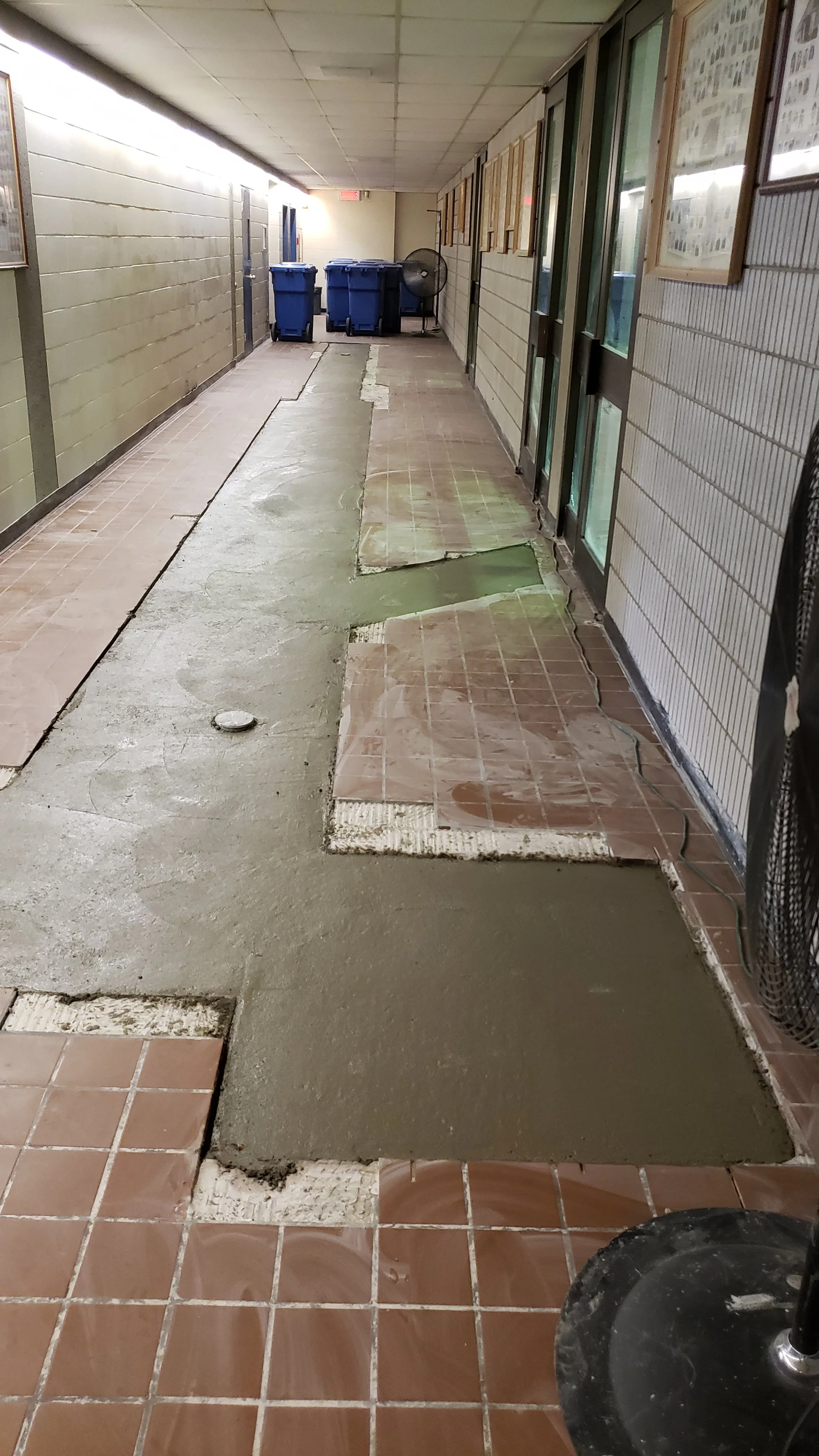 Rénovation d'un corridor avec un nouveau revêtement de sol en béton frais, en cours d'installation sur des carreaux de céramique existants, dans une école ou un bâtiment public, avec des poubelles bleues et un ventilateur en arrière-plan.