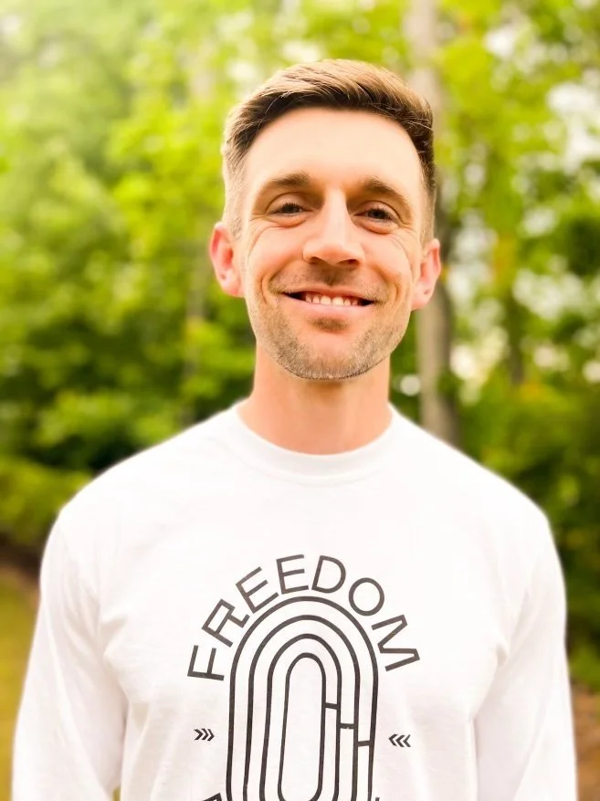 Man smiling outdoors wearing a white "FREEDOM" shirt, green trees in background.