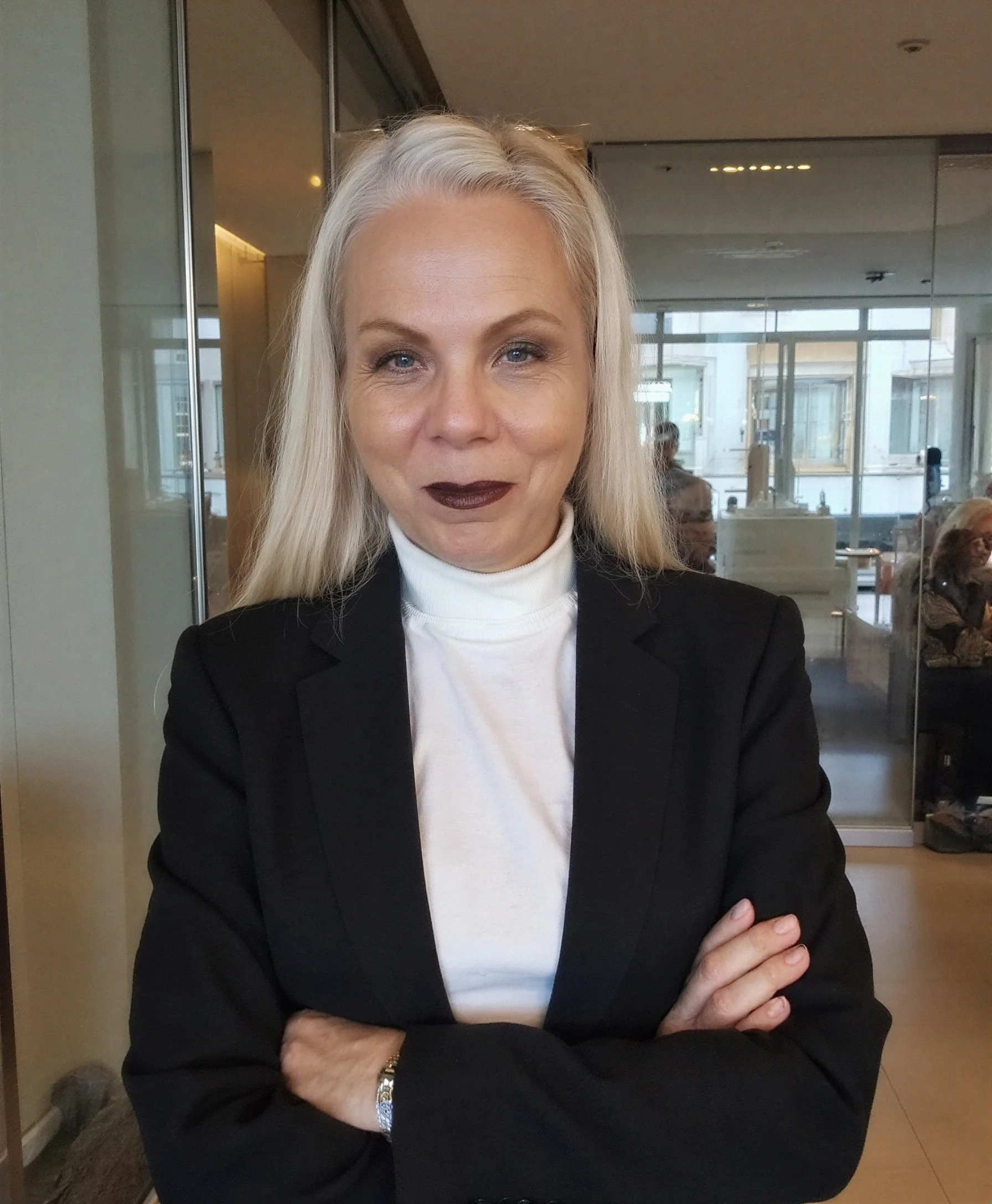 A woman with platinum blonde hair and dark lipstick posing with arms crossed inside a modern café or restaurant.