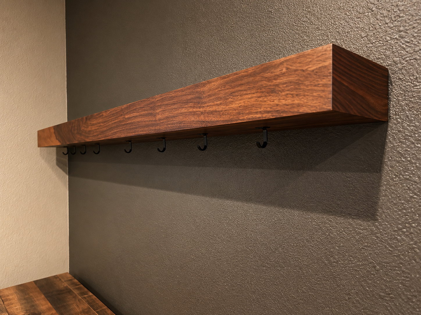 A wooden shelf mounted on a textured gray wall with black hooks mounted below the floating shelf.