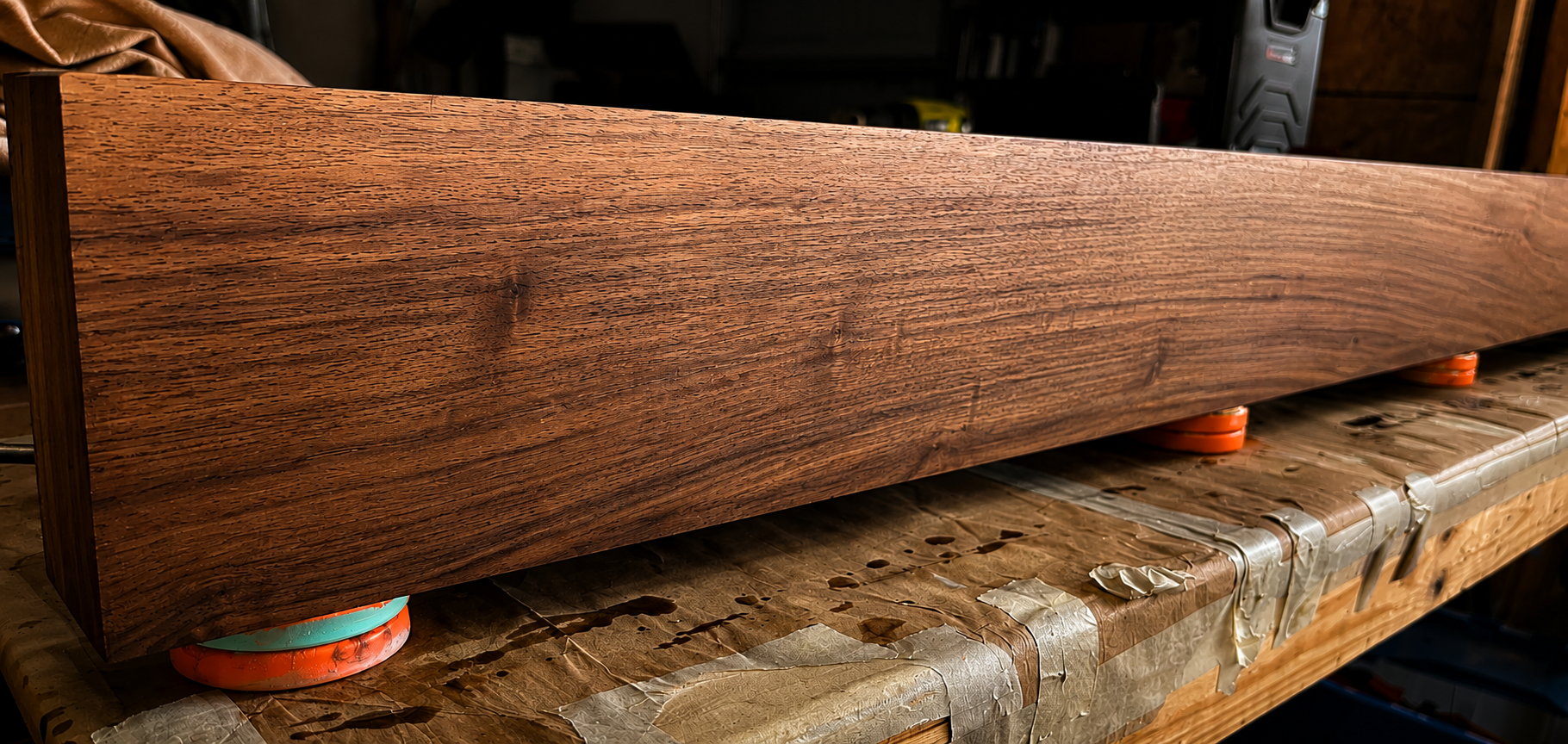 A long wooden beam with a rich, dark finish resting on orange and blue supports on a workbench covered with protective paper, in a woodworking shop.