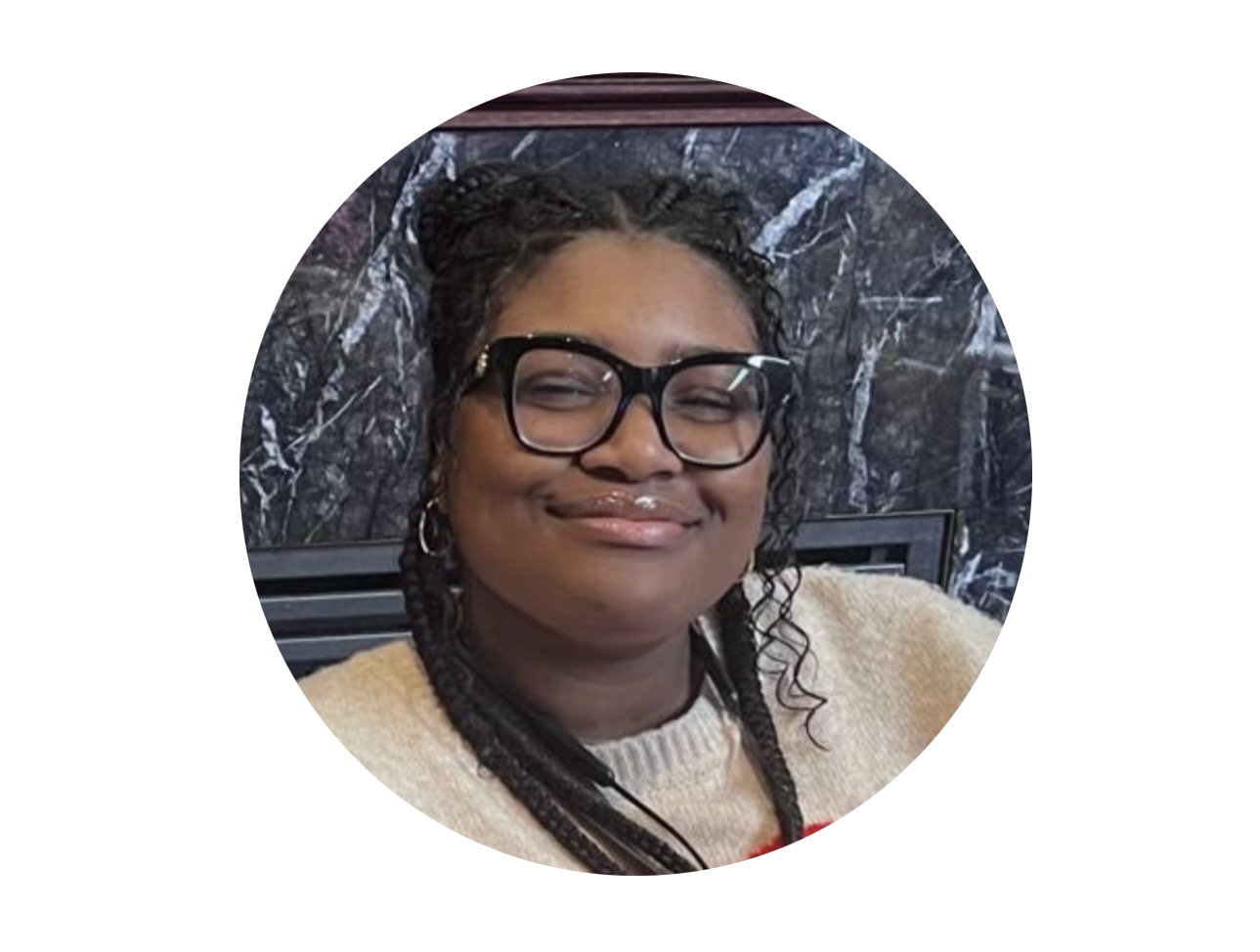 Smiling woman wearing glasses, earrings, and a light-colored sweater, sitting against a dark marble background.