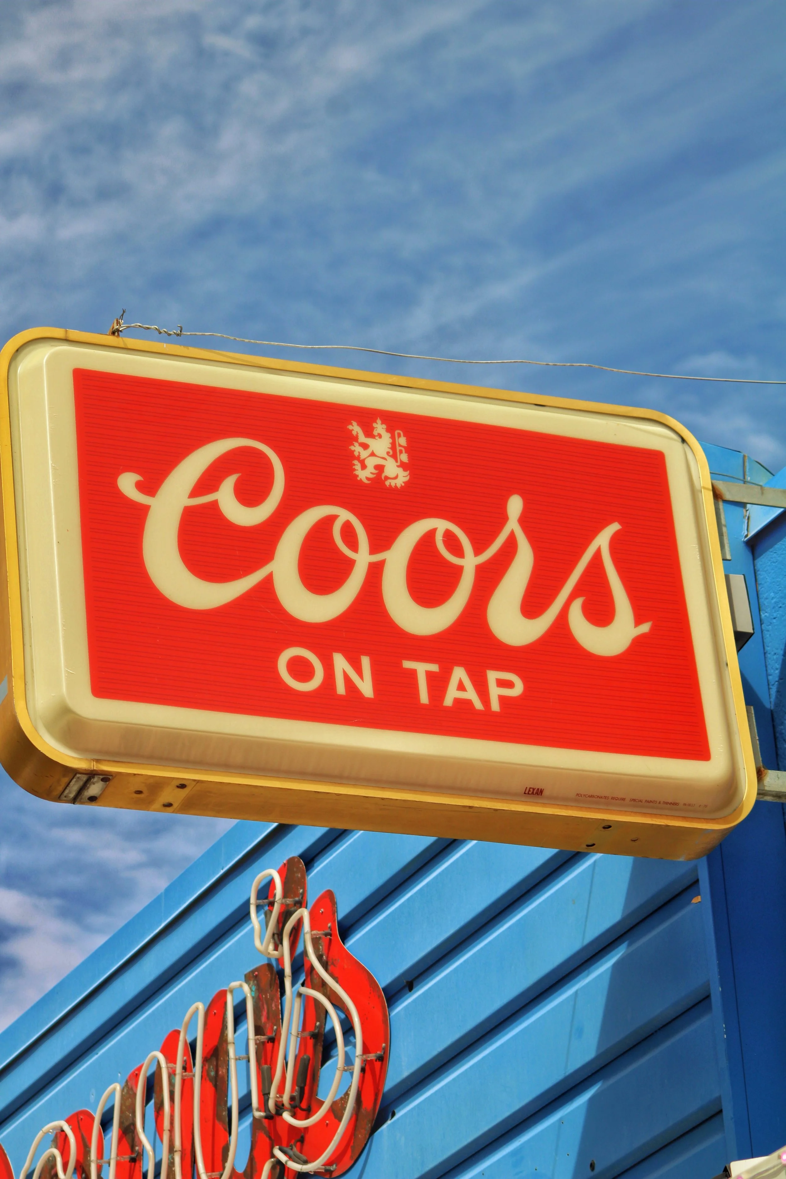 A bright red and cream sign reading 'Cools on Tap' hanging outside a blue building with a clear blue sky in the background.