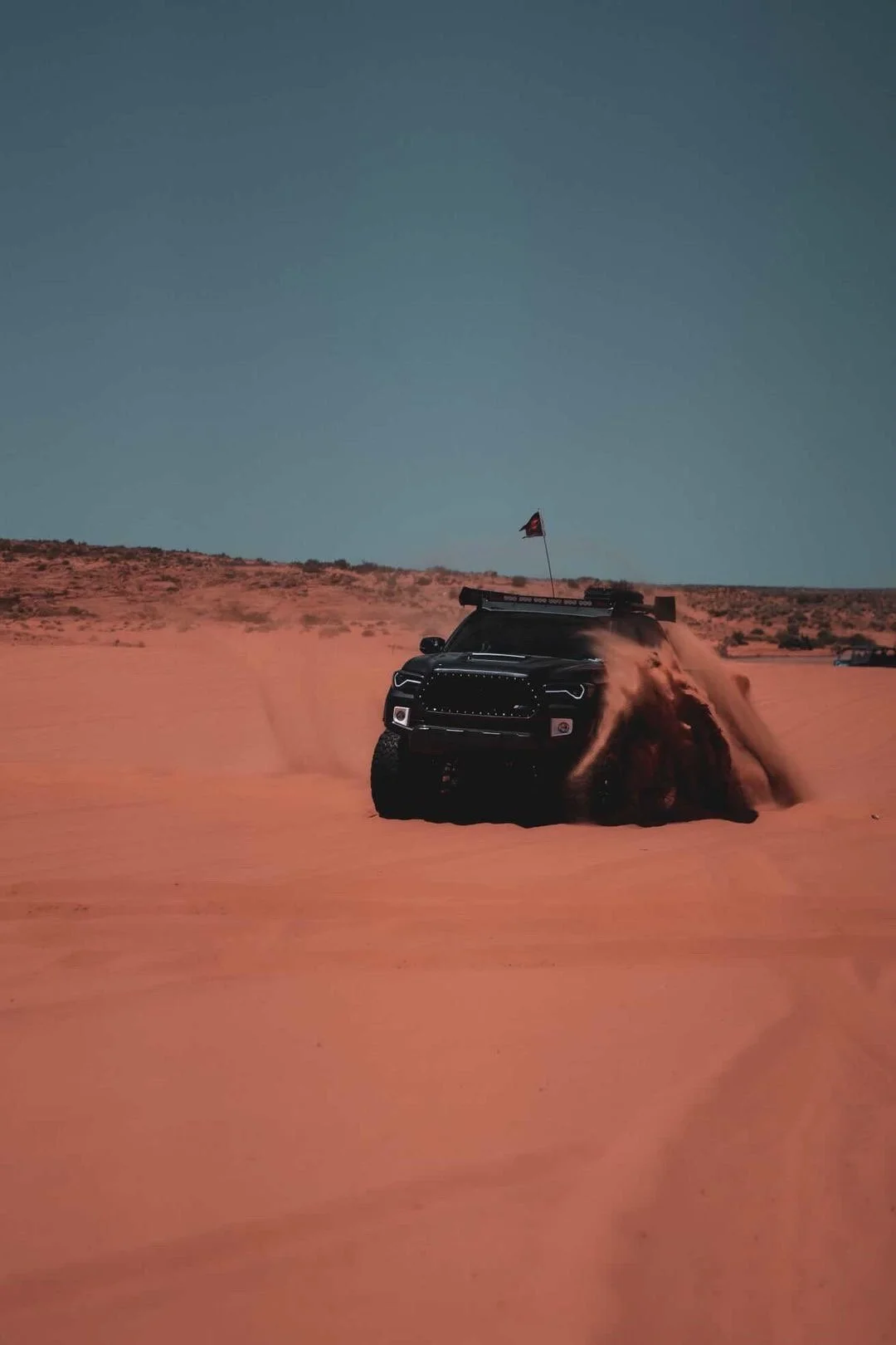 A black pickup truck kicking up sand in a desert landscape with orange sand dunes and a clear blue sky, flying a small flag on top.