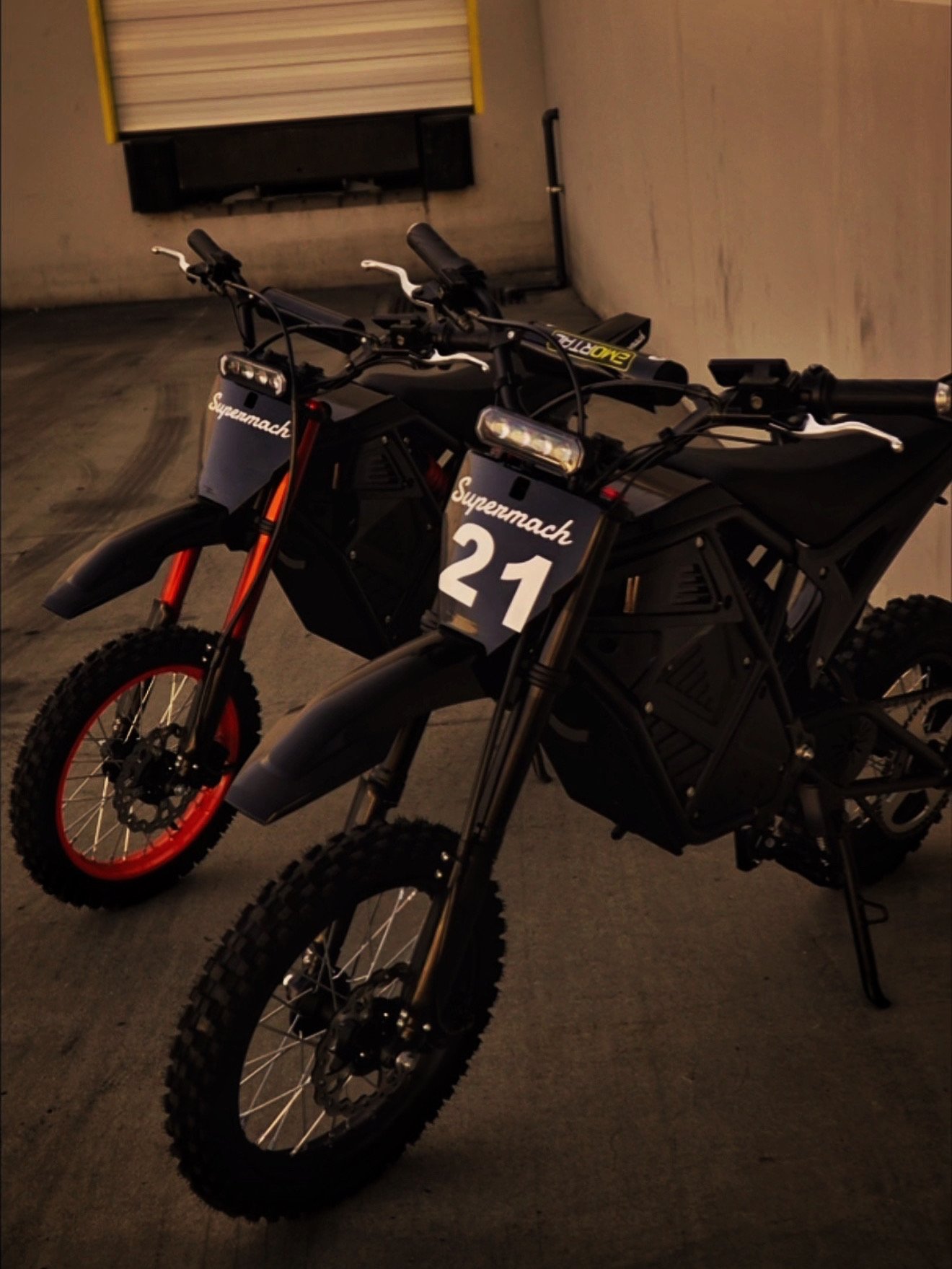 Two black electric dirt bikes with orange rims parked in an indoor space, labeled with 'Supermach 21' and 'Supermach' and equipped with headlamps.