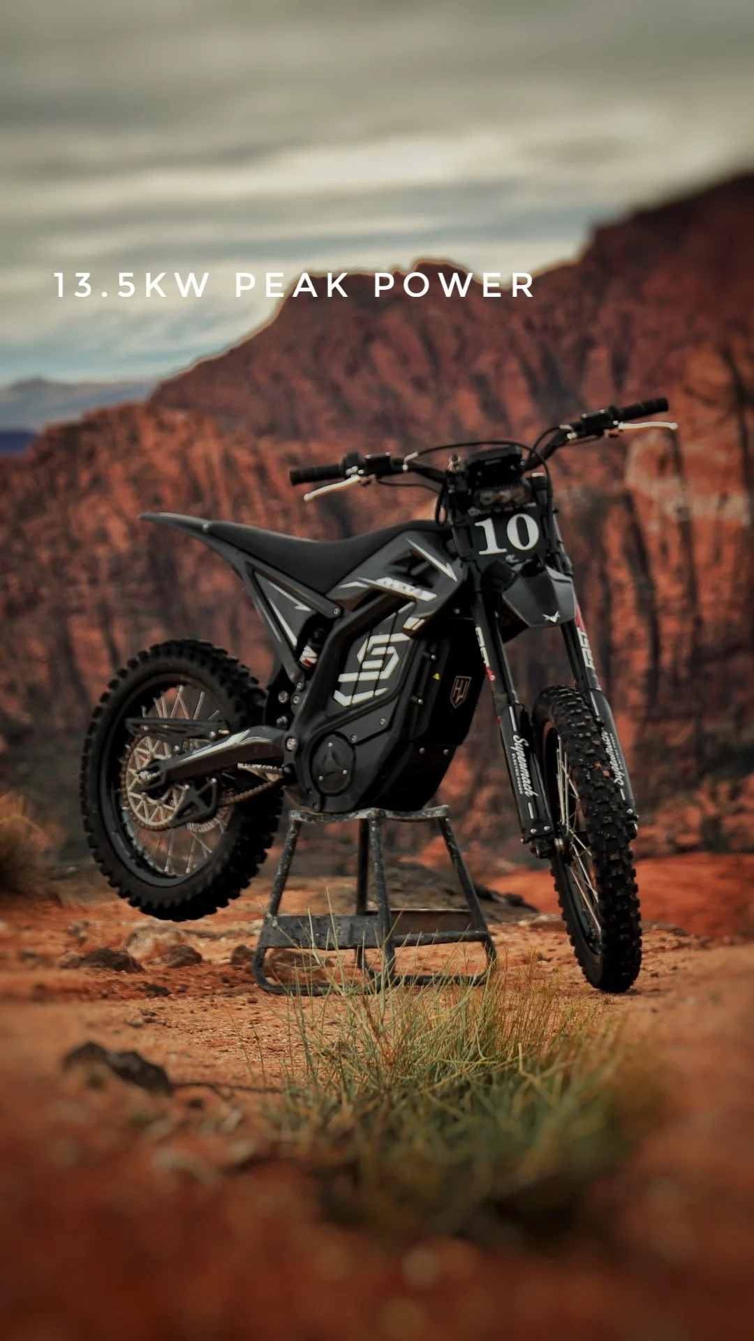 An electric dirt bike on a stand in front of a desert landscape with red rock formations, with the text '13.5KW PEAK POWER' overlaid.