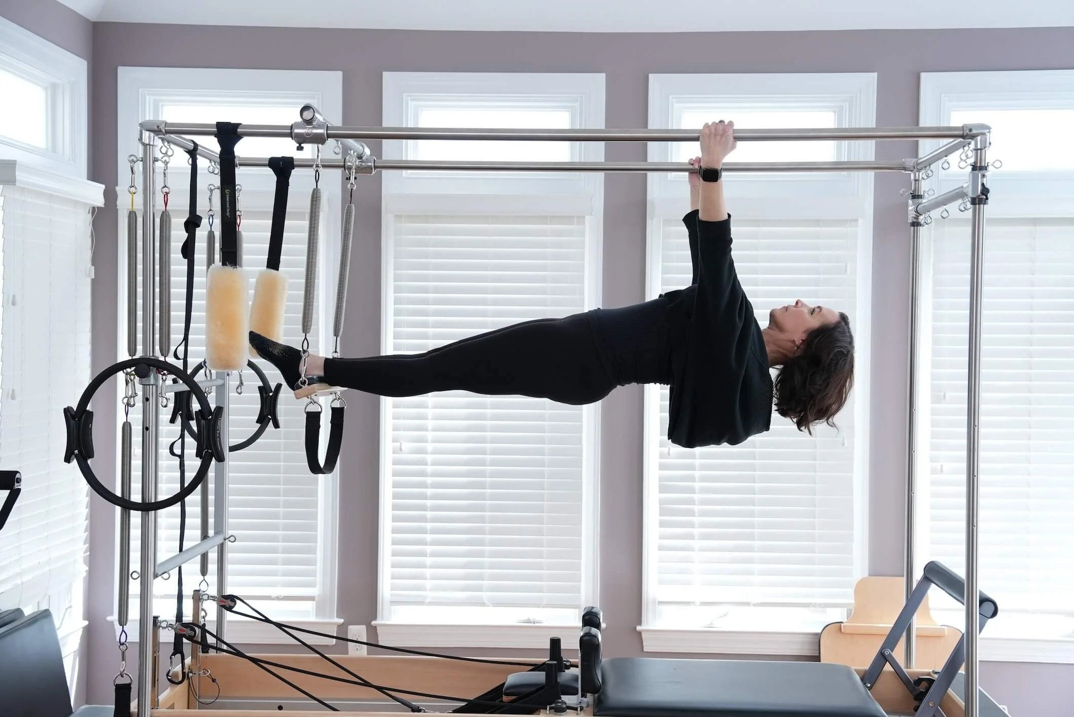 Woman exercising on a Pilates Cadillac reformer machine in a bright exercise room with large windows and white blinds.