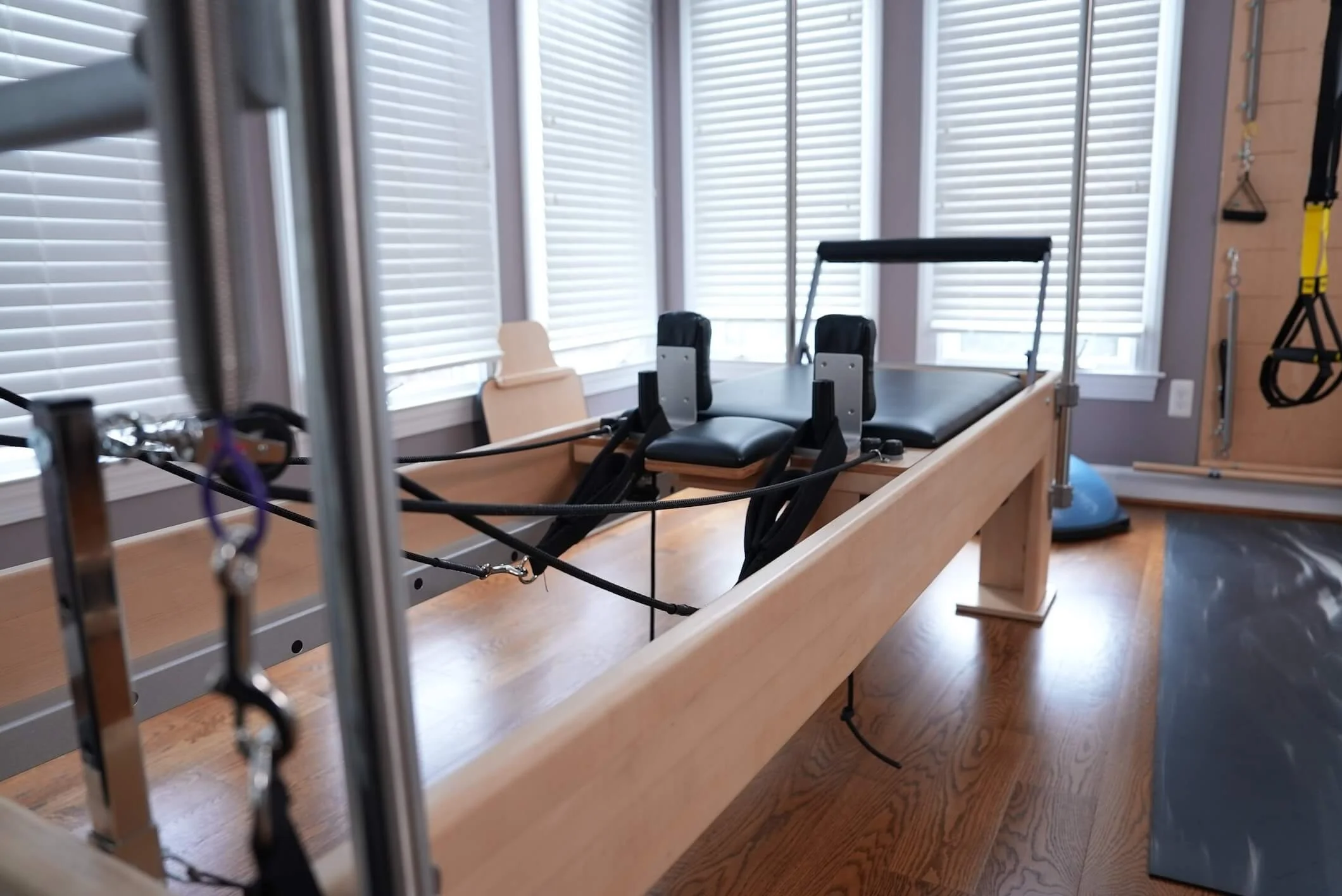 A Pilates reformer machine with a wooden frame and black padded supports in a room with wooden flooring and large windows with white blinds.