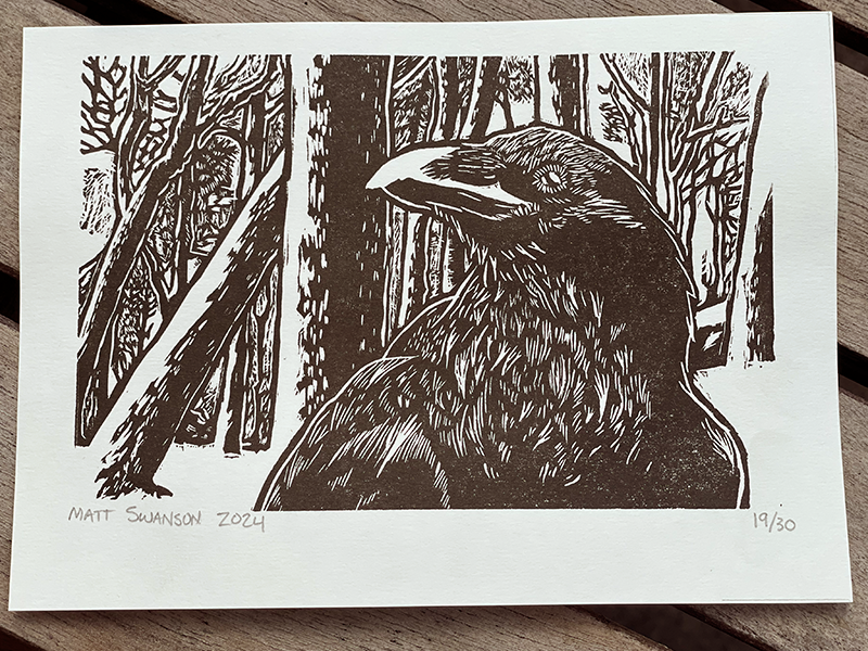 Photo of a black and white linocut art print featuring a raven in front of a snowy forest background, made by Matt Swanson