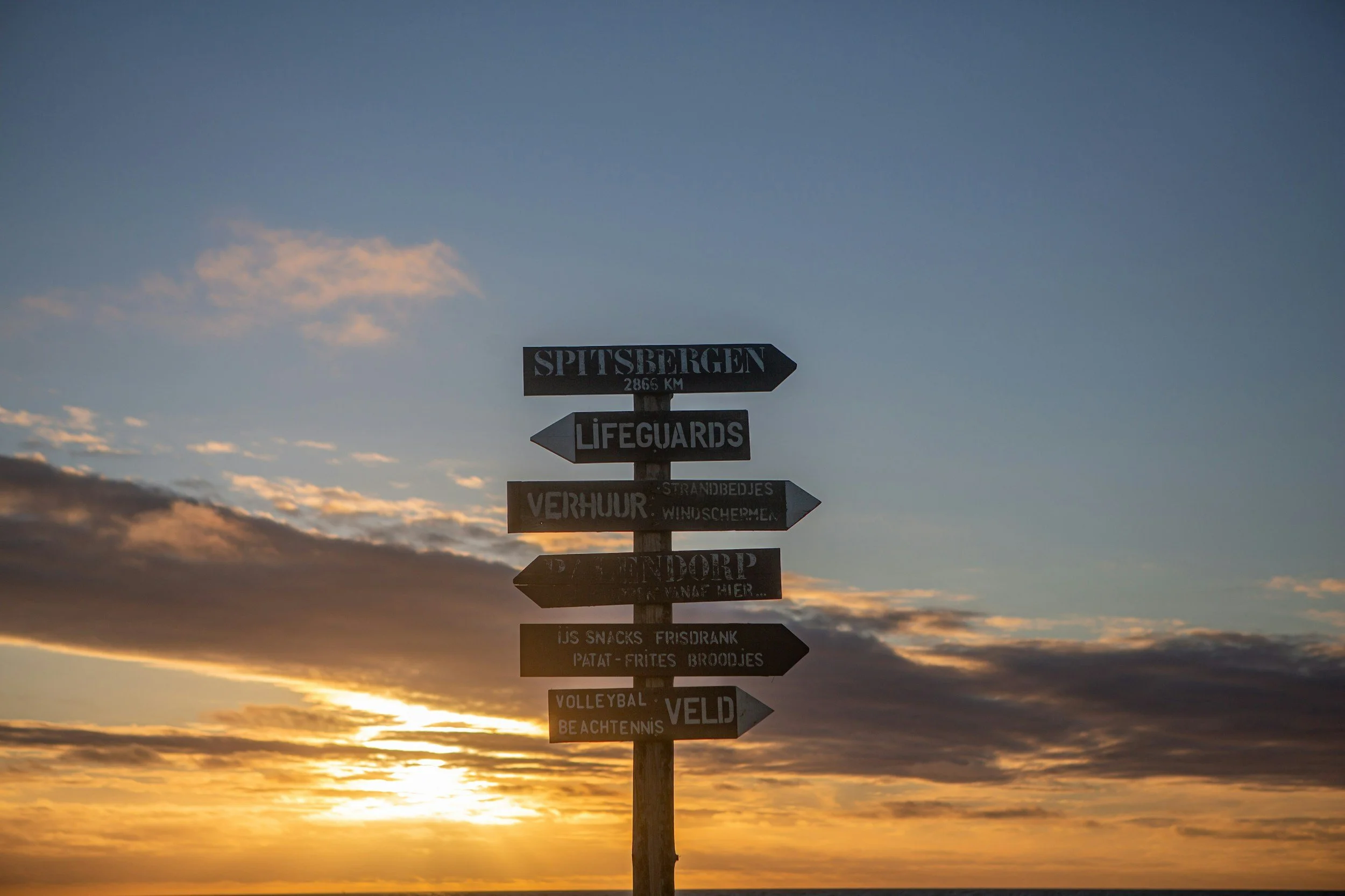 Signpost with multiple directional signs against a sunset sky at the beach, indicating directions to Spitsbergen, lifeguards, rental, endorp, and beach activities.